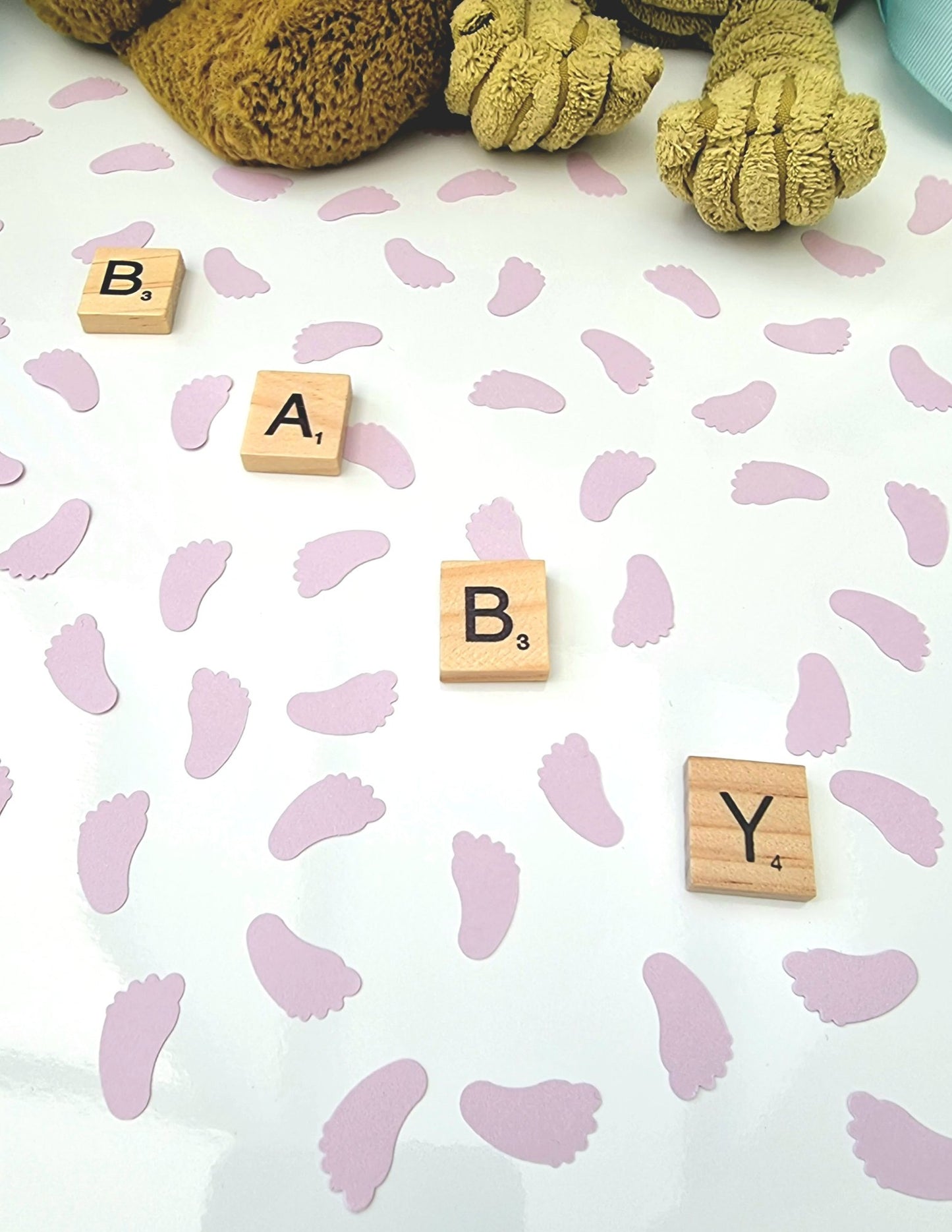 Pastel lilac baby feet shaped confetti pieces are scattered on a white background with wooden scrabble tiles reading BABY diagonally across the photo. Cuddly toys are seen in the background