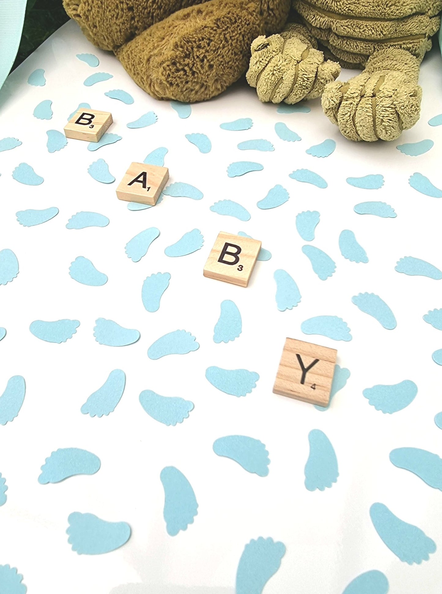 Pastel blue baby feet shaped confetti pieces are scattered on a white background with wooden scrabble tiles reading BABY diagonally across the photo. Cuddly toys are seen in the background