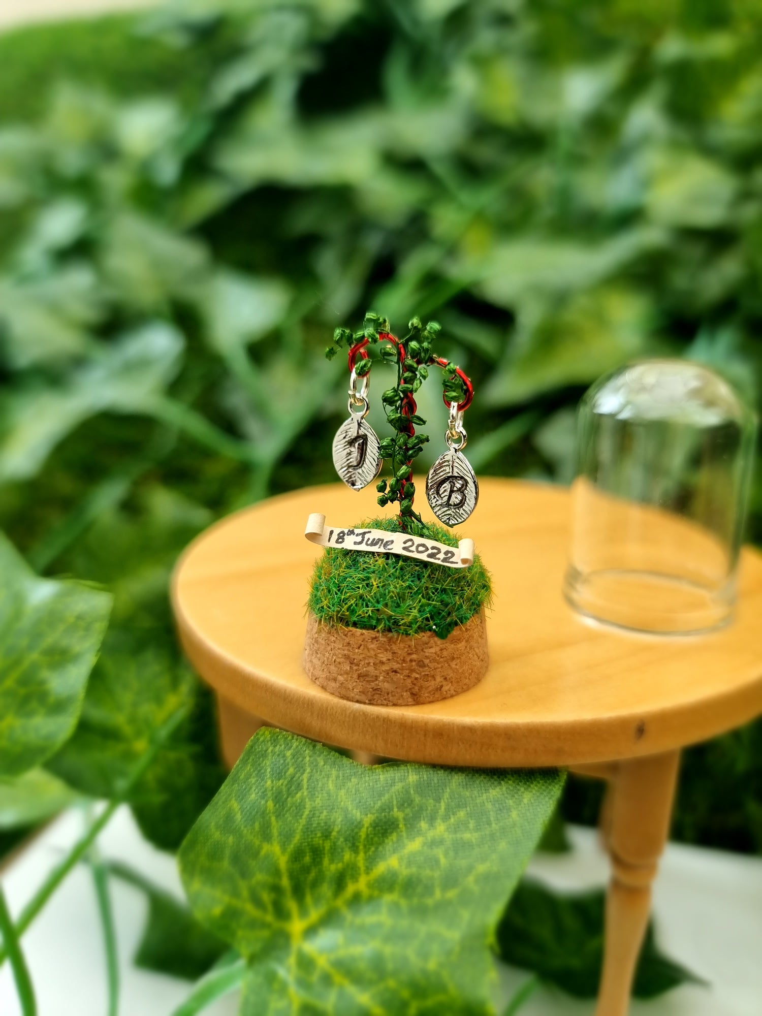 Miniature diorama sits on a 12th scale table. On top of the cork base is a small grassy hill with a red wire crook twisted with green foliage. 2 leaf shape monogram charms hang from the crook above the grass and above a tiny scroll with the date of the wedding.