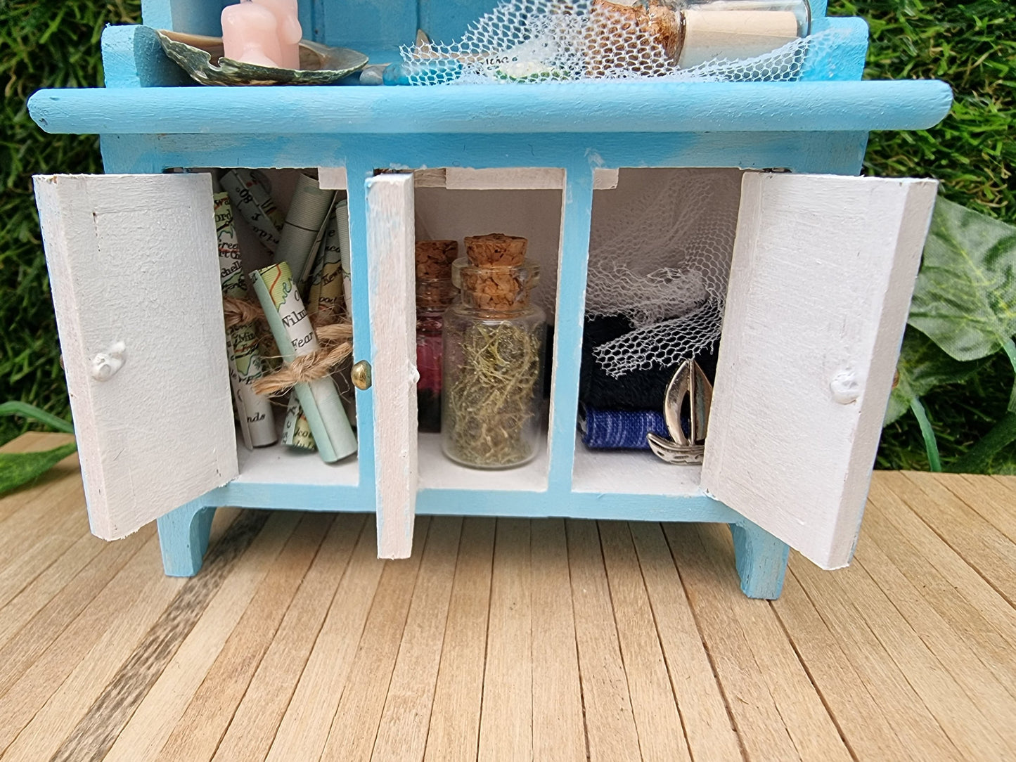 A 1:12 miniature pastel blue dresser sits on a wooden floor. The dresser had 3 open cupboard doors, revealing a silver coloured ship, maps and bottles inside