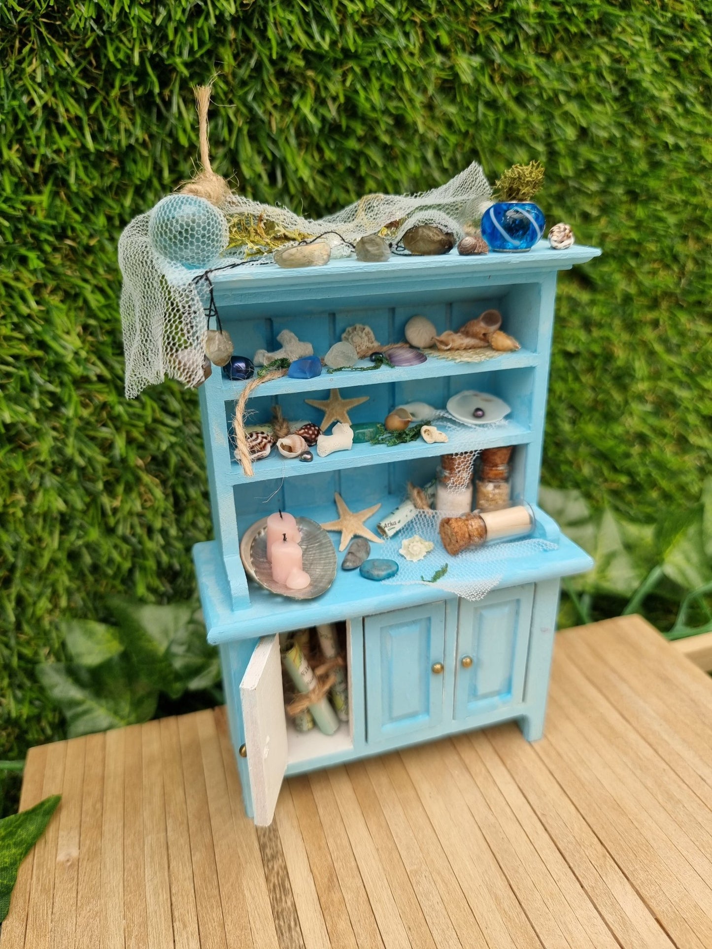 A 1:12 miniature pastel blue dresser sits on a wooden floor against a green background. The dresser displays glass vials, netting, glass floats, shells and candles in a shell dish. The dresser had 3 cupboard doors. One is open revealing maps inside