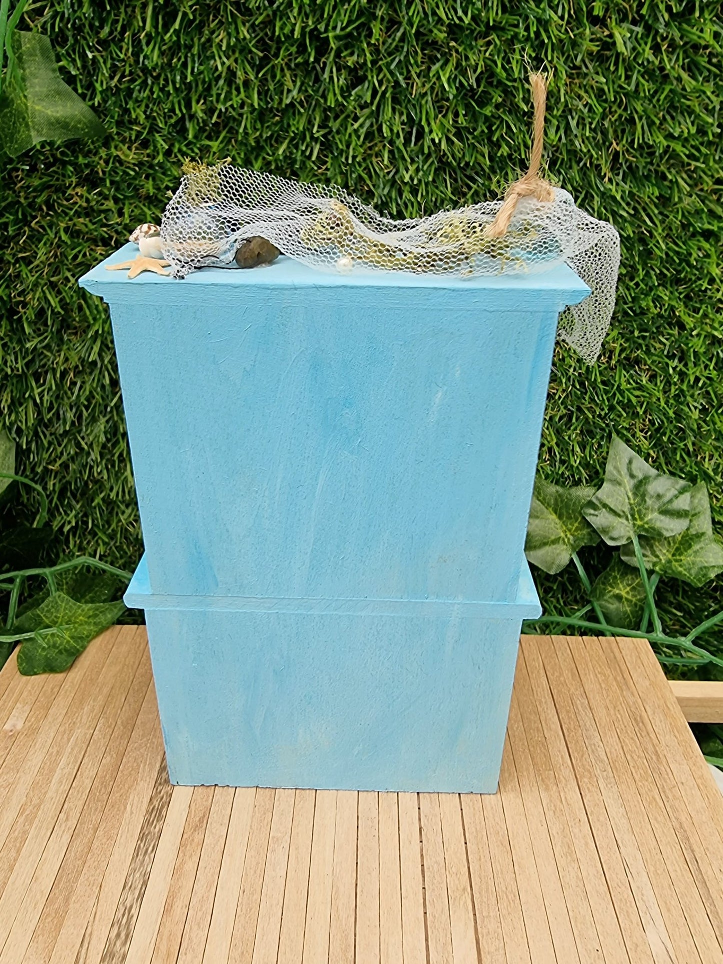 The back of a 1:12 scale miniature pastel blue dresser on a wooden floor against a green background. Netting and glass floats can be seen on top