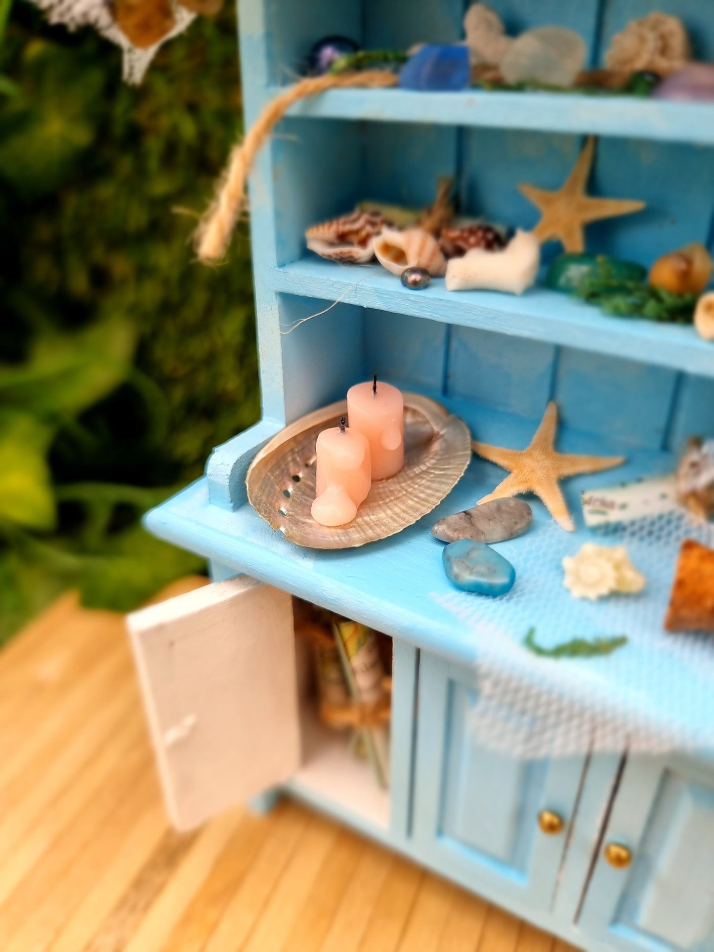 A 1:12 miniature pastel blue dresser sits on a wooden floor against a green background. The dresser displays glass vials, netting, glass floats, shells and candles in a shell dish. The dresser had 3 cupboard doors. One is open revealing maps inside