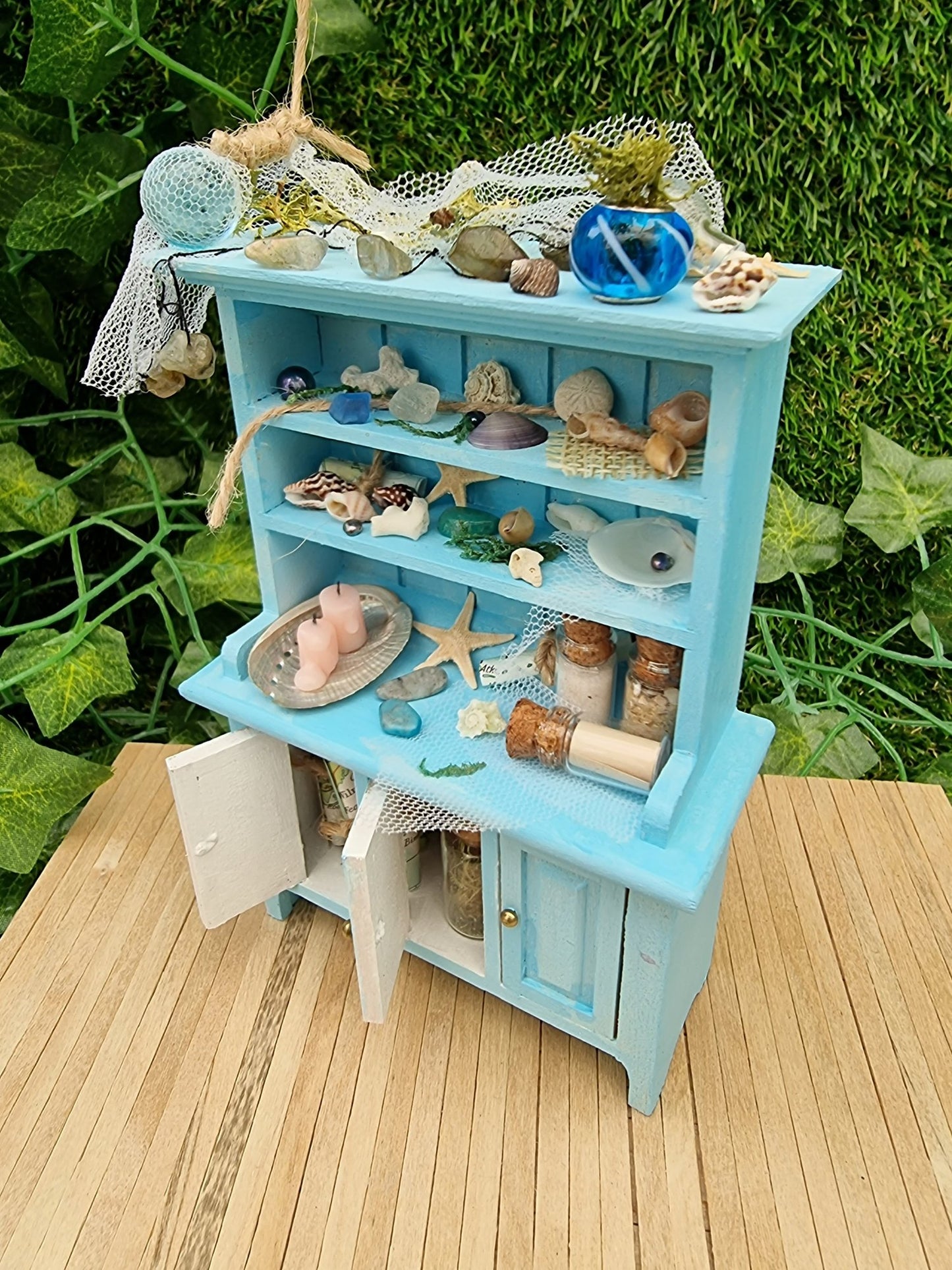 A 1:12 miniature pastel blue dresser sits on a wooden floor against a green background. The dresser displays glass vials, netting, glass floats, shells and candles in a shell dish. The dresser had 3 cupboard doors. Two are open revealing maps inside