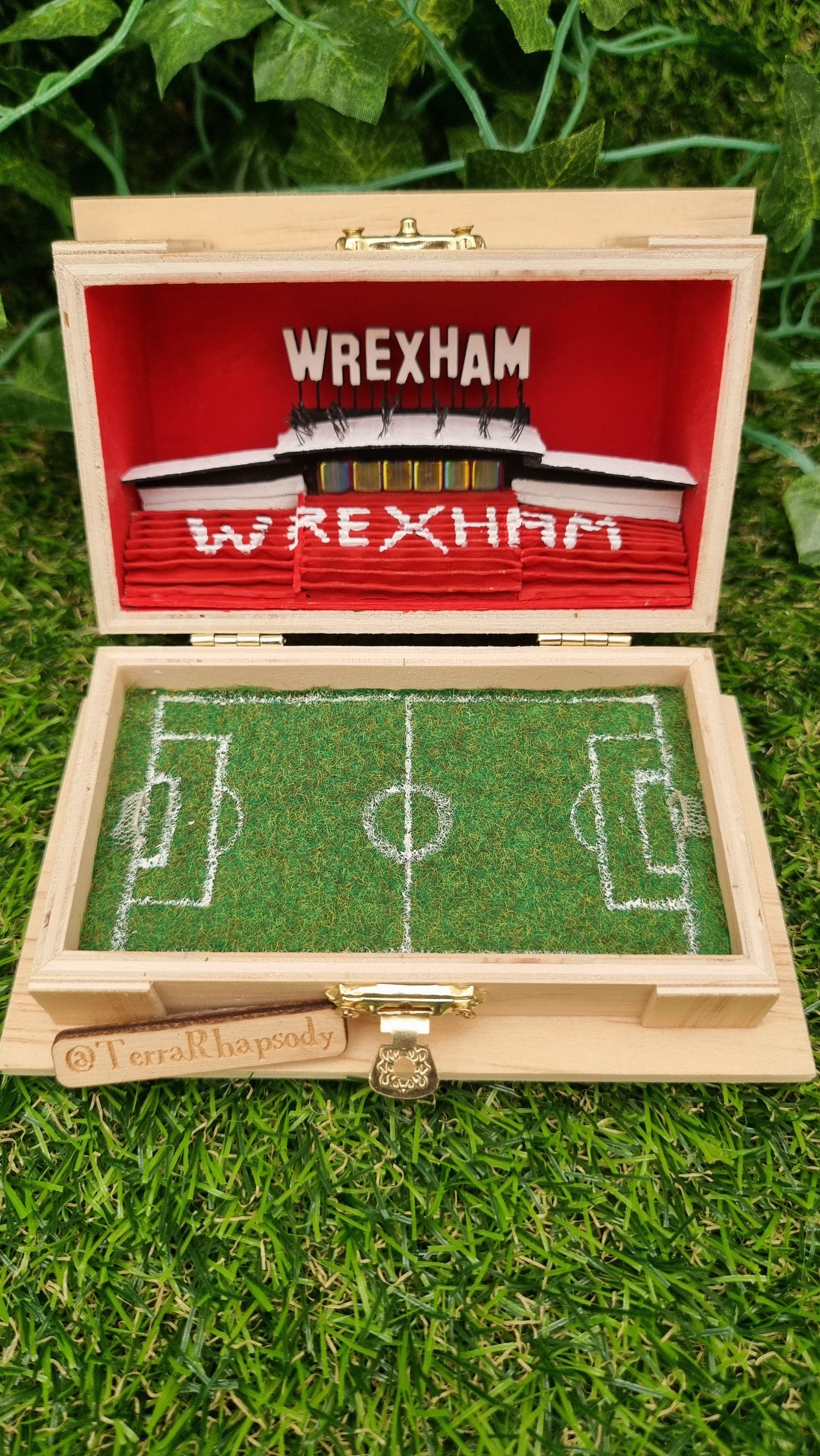 A small wooden box sits open with a miniature diorama of Wrexham AFC stadium inside, including stands, executive boxes and the pitch with tiny goals