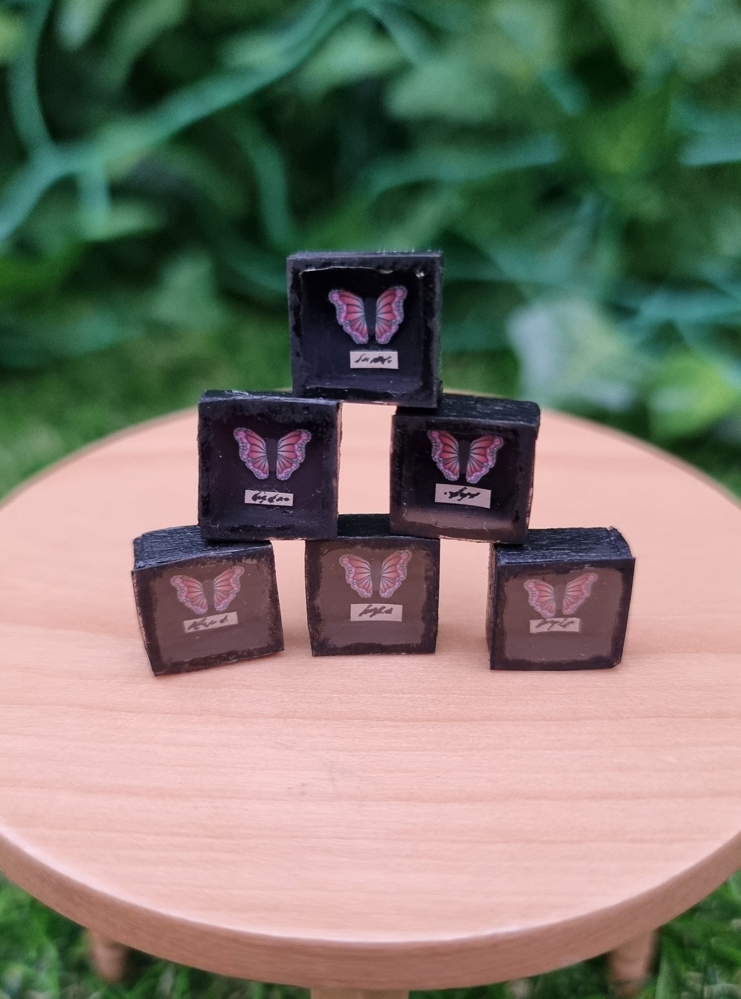 6 1:12 scale miniature shadow boxes containing pink and purple butterflies are stacked up on a miniature wooden table against a green background