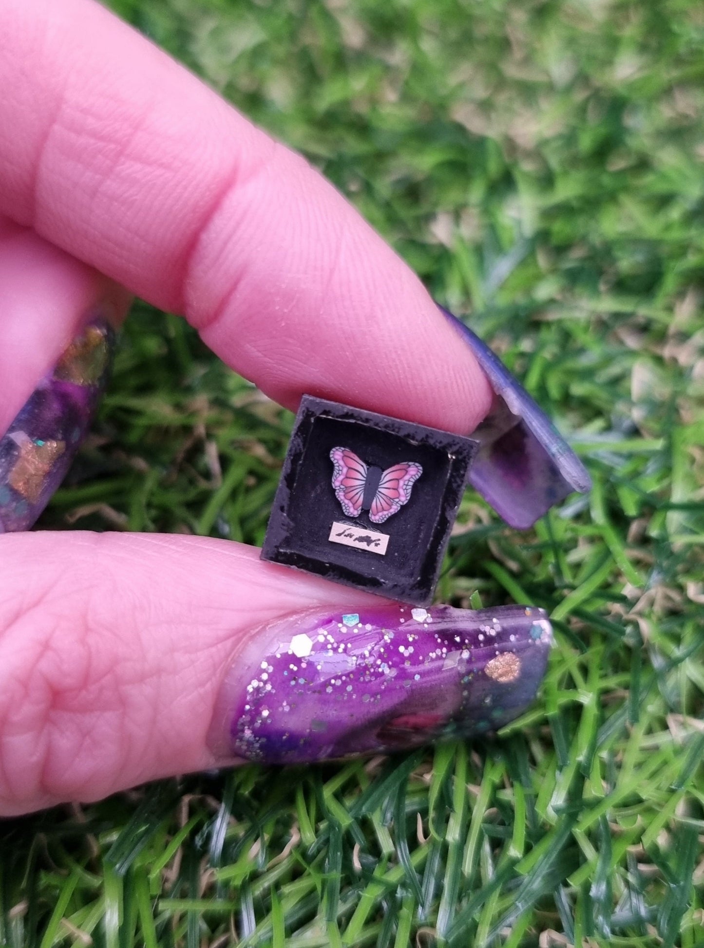 1:12 scale black shadow box containing a pink and purple butterfly is held in a hand. There is a tiny strip of paper beneath the butterfly with indistiguishable text. Displayed against green grass background