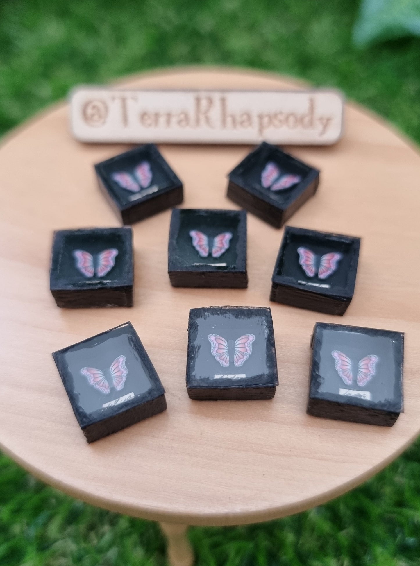 8 1:12 scale miniature black shadow boxes containing pink and purple butterflies are displayed on a miniature wooden table with a sign in the background with @TerraRhapsody