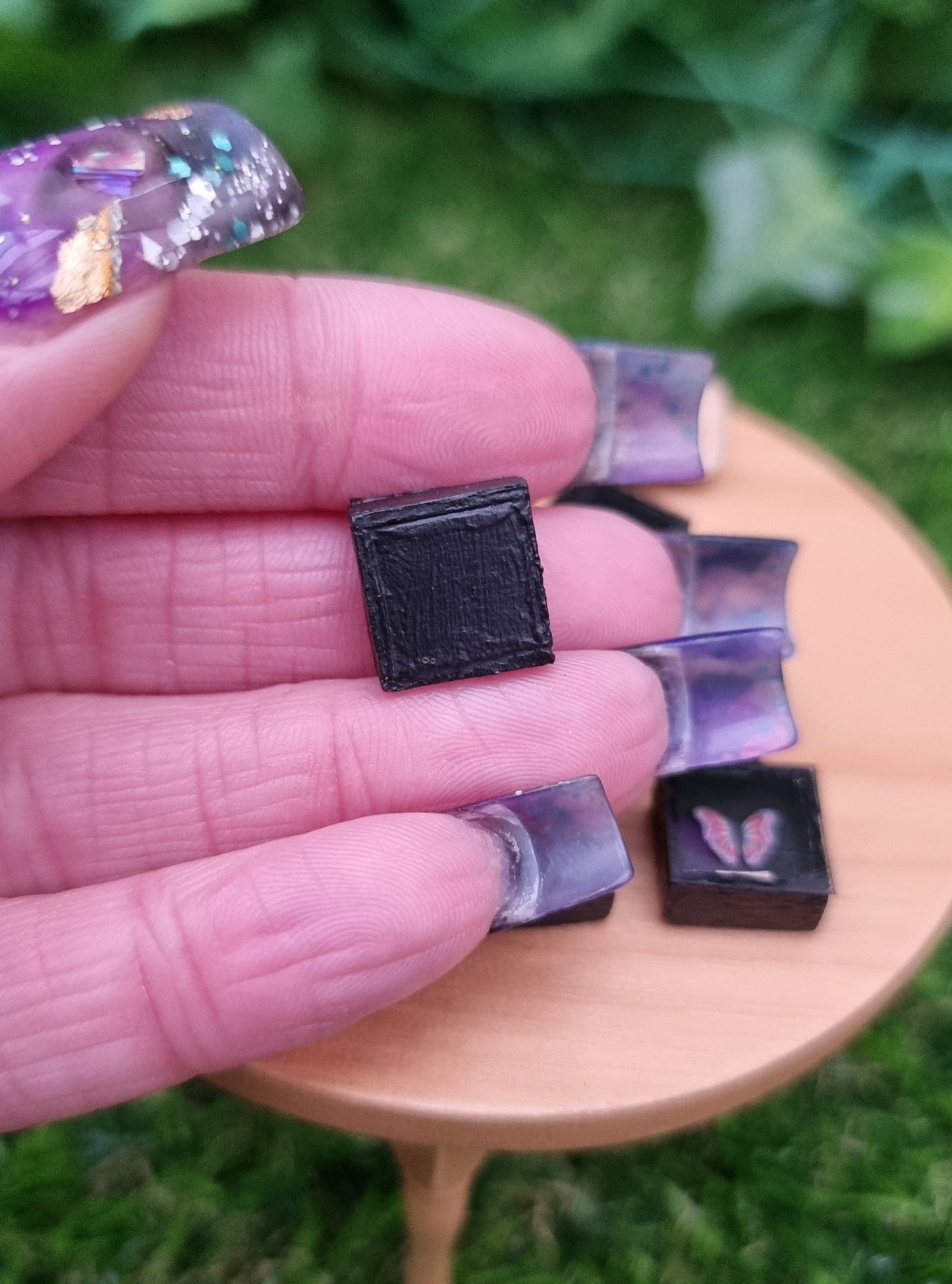 A hand holds a 1:12 scale miniature black shadow box. The back of the box is shown. More shadow boxes with pink and purple butterfly is shown in the background.