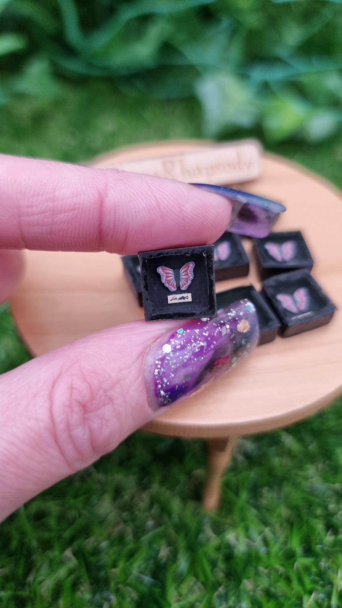 1:12 scale black shadow box containing a pink and purple butterfly is held in a hand. There is a tiny strip of paper beneath the butterfly with indistiguishable text. More butterfly shadow boxes can be seen in the background