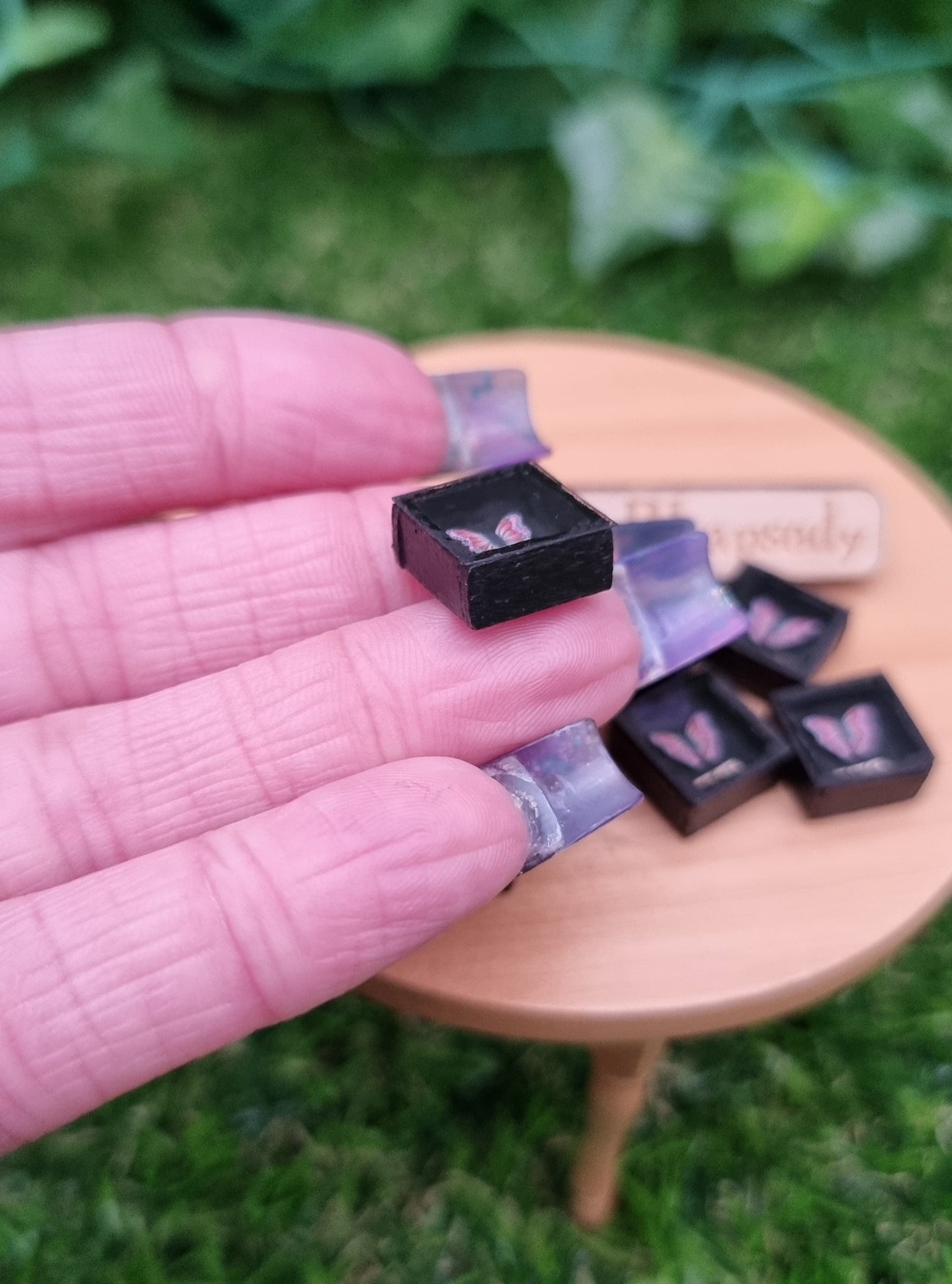 A hand holds a 1:12 scale miniature black shadow box. The side of the box is shown with the butterfly visible inside. More shadow boxes with pink and purple butterflies is shown in the background.