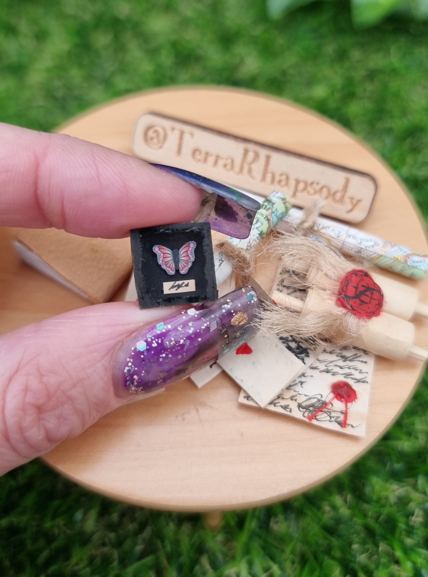 1:12 scale black shadow box containing a pink and purple butterfly. There is a tiny strip of paper beneath the butterfly with indistiguishable text. Map rolls, letters and parchments can be seen in the background