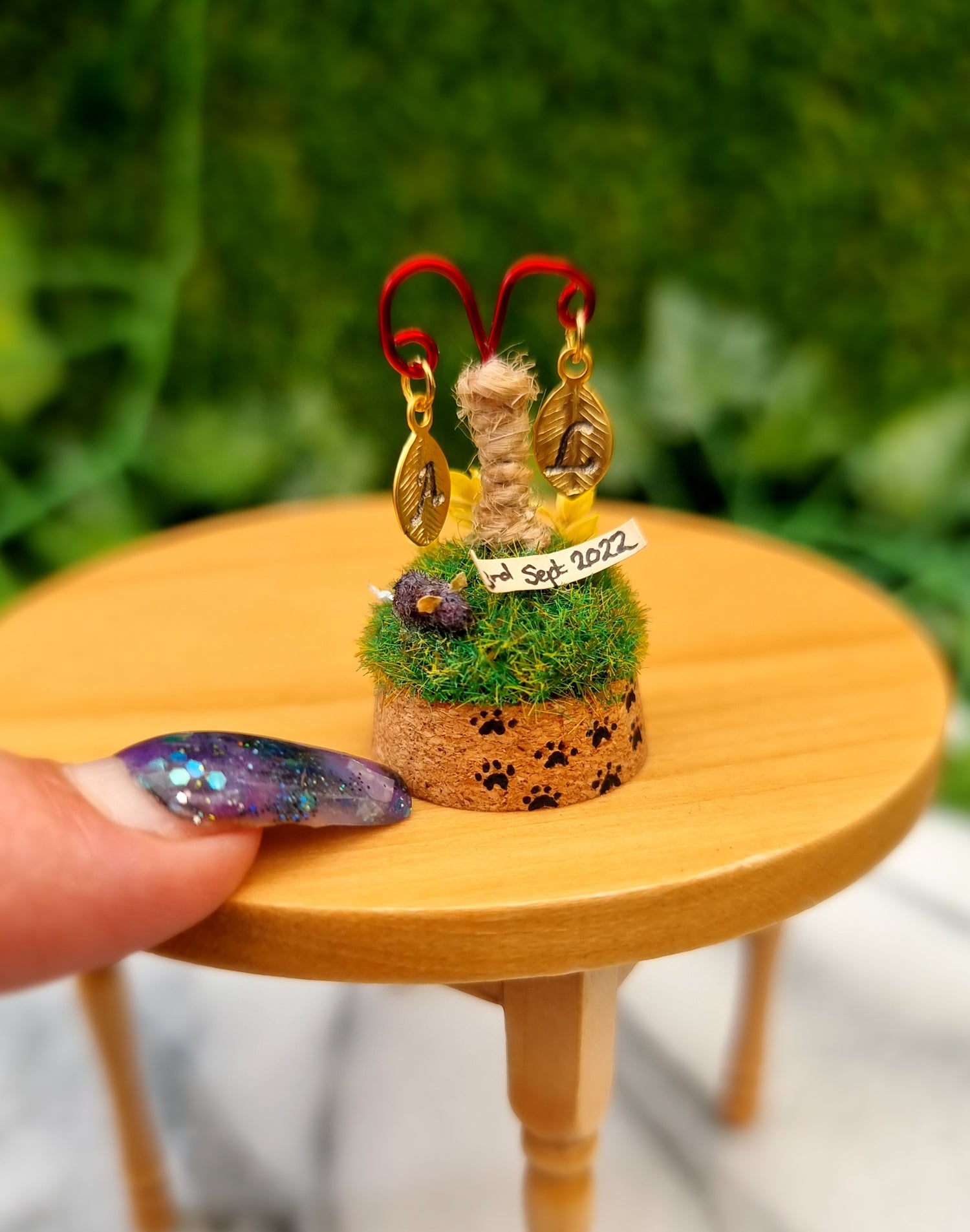 Miniature diorama sits on a 12th scale table. It has a cork base with a trail of tiny paw prints. On top is a small grassy hill with a cat scratch post topped with a wire crook. 2 leaf shape monogram charms hang from the crook above a tiny mouse toy and dated slip of paper on the grass. A finger points for scale
