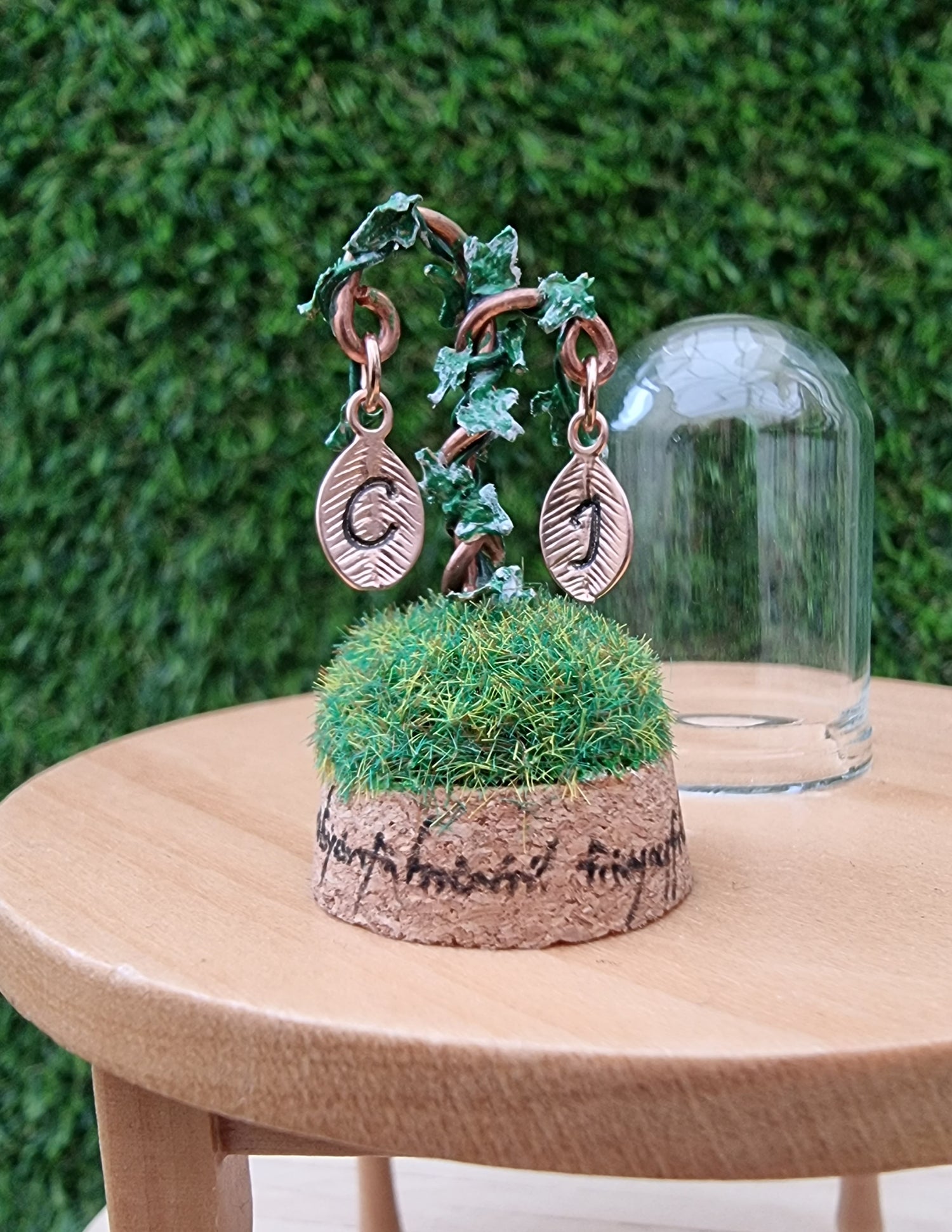 Miniature diorama sits on a 12th scale table. Elvish text surrounds the cork base. On top is a small grassy hill with a copper wire crook twisted with green and white variegated ivy. 2 leaf shape monogram charms hang from the crook above the grass