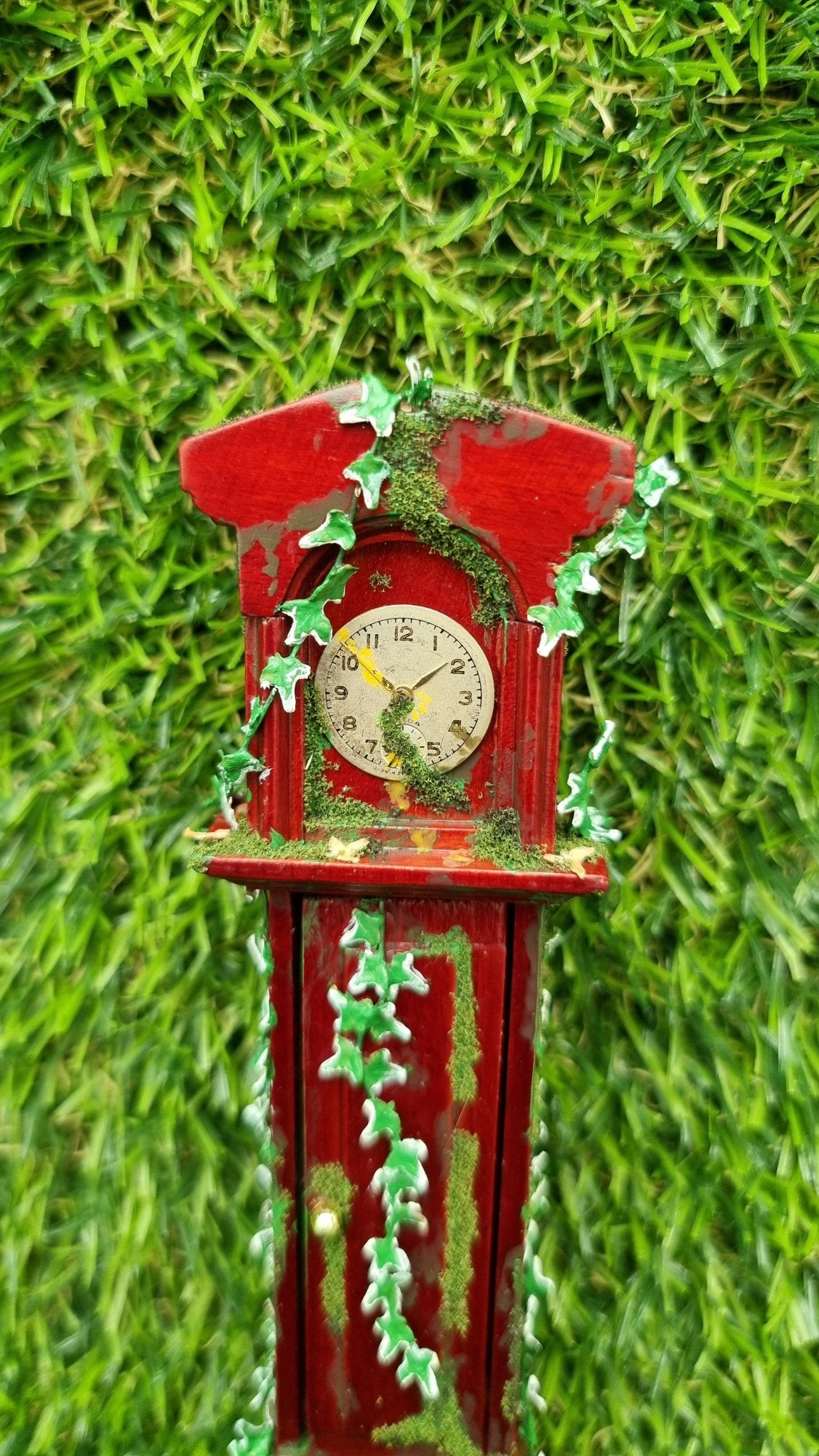 Miniature 1:12 scale grandfather clock sits against a green background. The top half of the clock is shown covered with climbing green ivy and moss. The clock face is stopped at 1.50