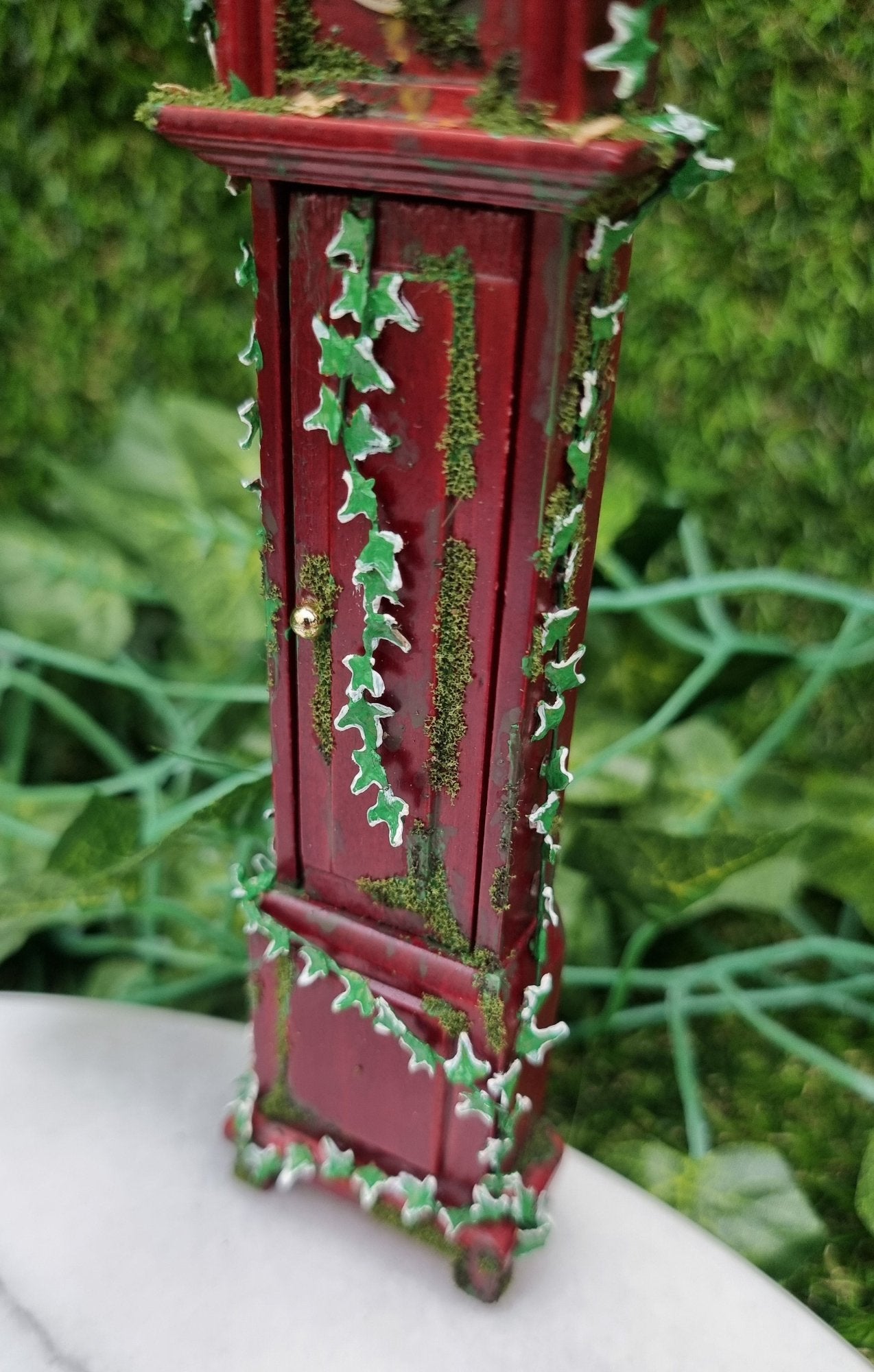 Miniature 1:12 scale grandfather clock sits of a marble slab, covered with climbing green ivy and moss. The clock face cannot be seen. The closed door in the body of the clock has ivy climbing down it