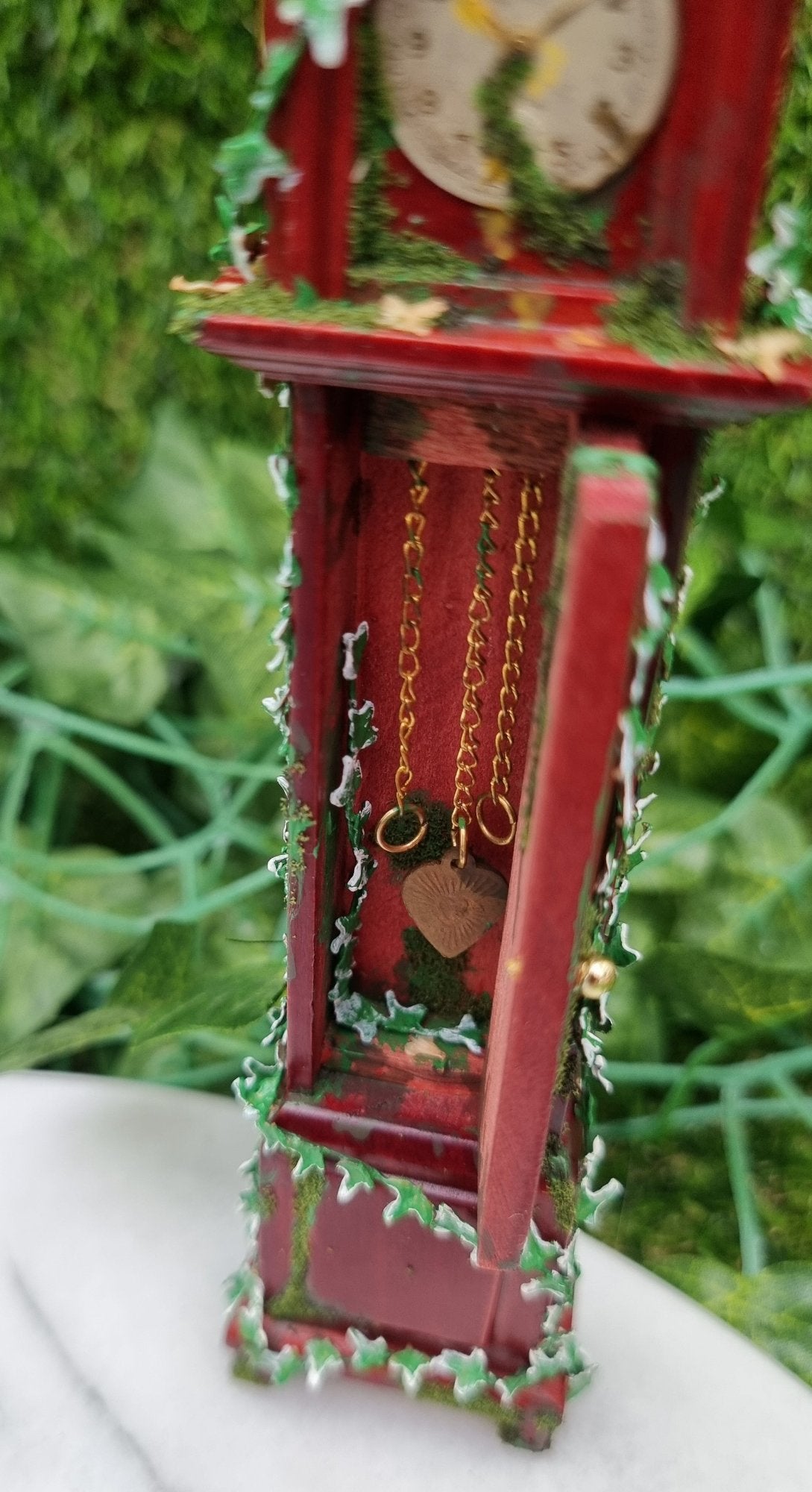 Miniature 1:12 scale grandfather clock sits of a marble slab, covered with climbing green ivy and moss. The clock face is stopped at 1.50 and the door in the body of the clock is open showing more ivy and hanging chains inside