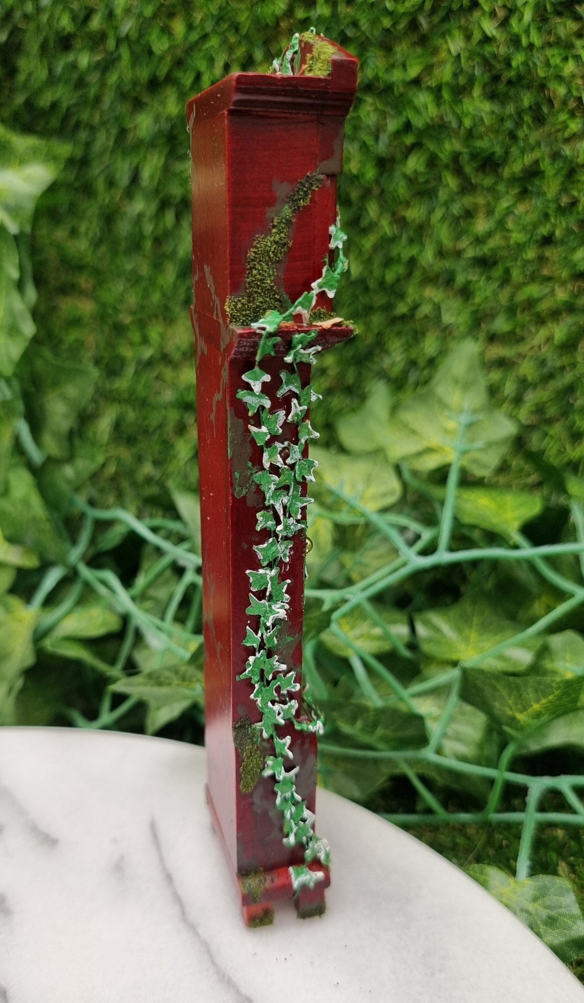 Miniature 1:12 scale grandfather clock sits of a marble slab, covered with climbing green ivy and moss. The side of the clock is shown, thick with green ivy with white variegation