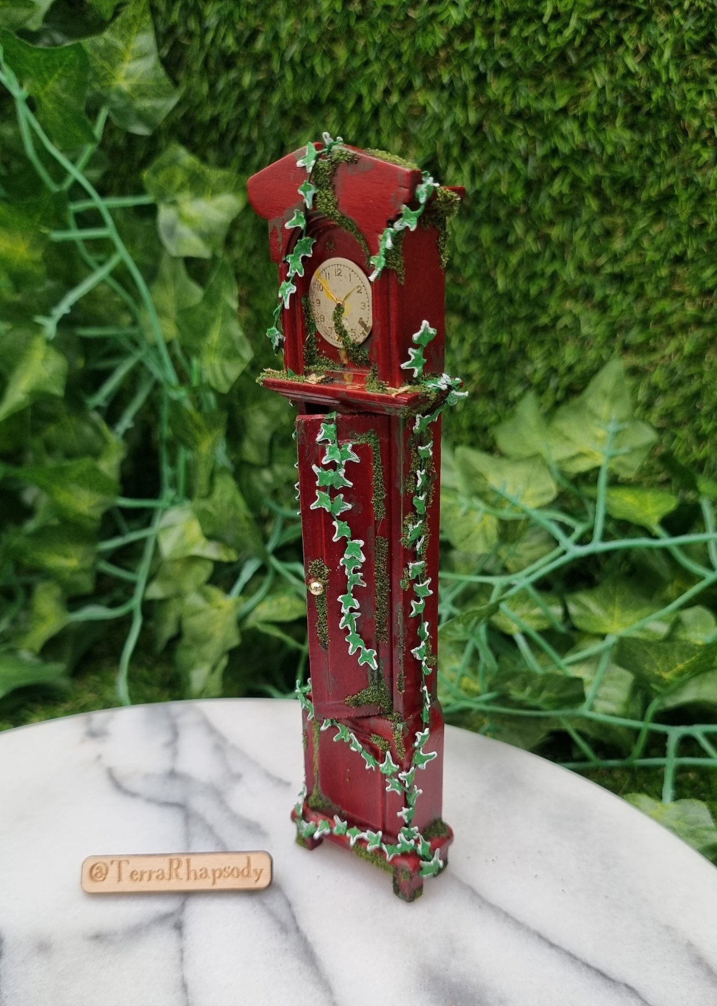 Miniature 1:12 scale grandfather clock sits of a marble slab, covered with climbing green ivy and moss. The clock face is stopped at 1.50 and the door in the body of the clock is ajar. @TerraRhapsody is written on wood beside it