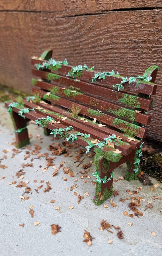 1:12 scale dollhouse wooden bench sits abandoned against a wood and stone background. The bench has green ivy twisting through the slats, dead leaves, grass and moss