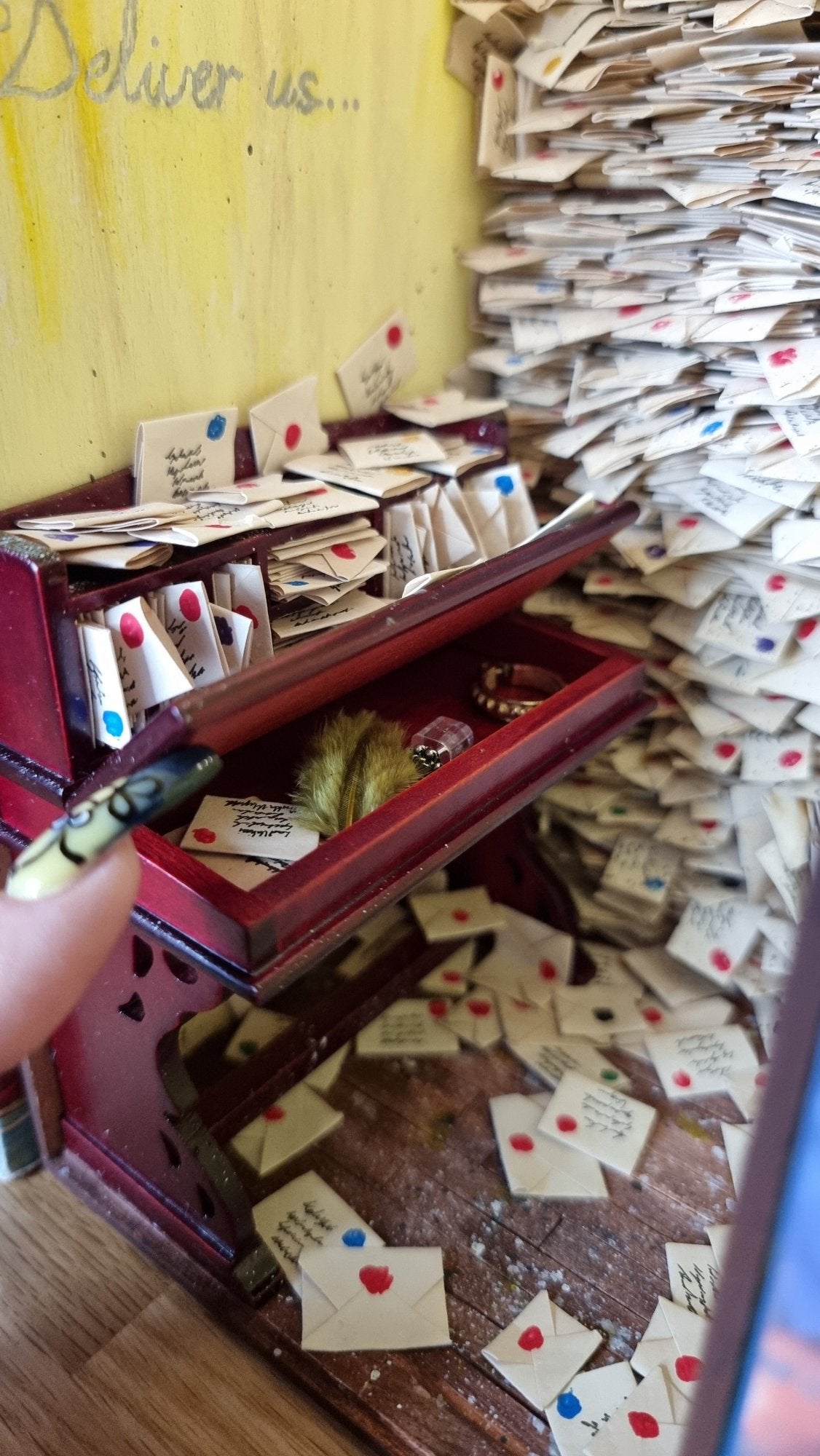 A finger points to a 1:12 scale miniature desk covered in letters. It sits against a yellow wall with silver cursive text reading "Deliver Us" above. In the background, shelves crammed with letters. The desk top is open revealing letters, quill and inkwell inside