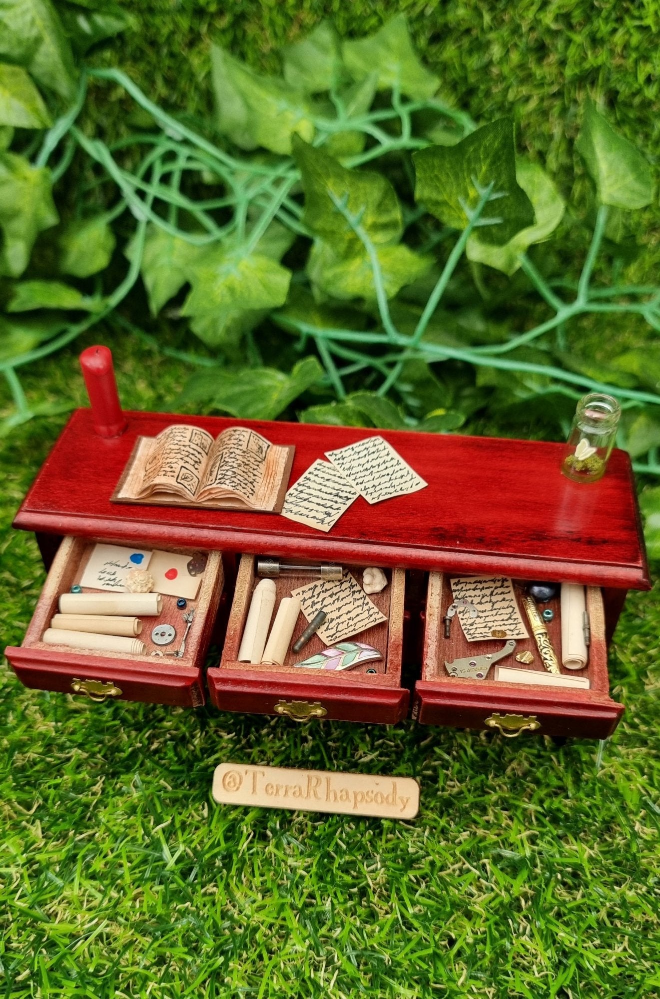 Miniature 1:12 scale dollhouse sideboard seen from above with 3 open drawers containing letters, parchments, trinkets and curios. The countertop has a red candle, open book, butterfly bottle and parchments