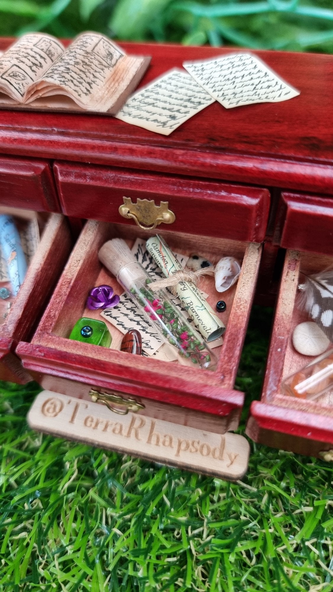 Miniature 1:12 scale dollhouse sideboard showing an open drawer containing bottle, map, parchment, trinkets and curios. The countertop has an open book and parchments. @TerraRhapsody is written on a piece of wood beside it.