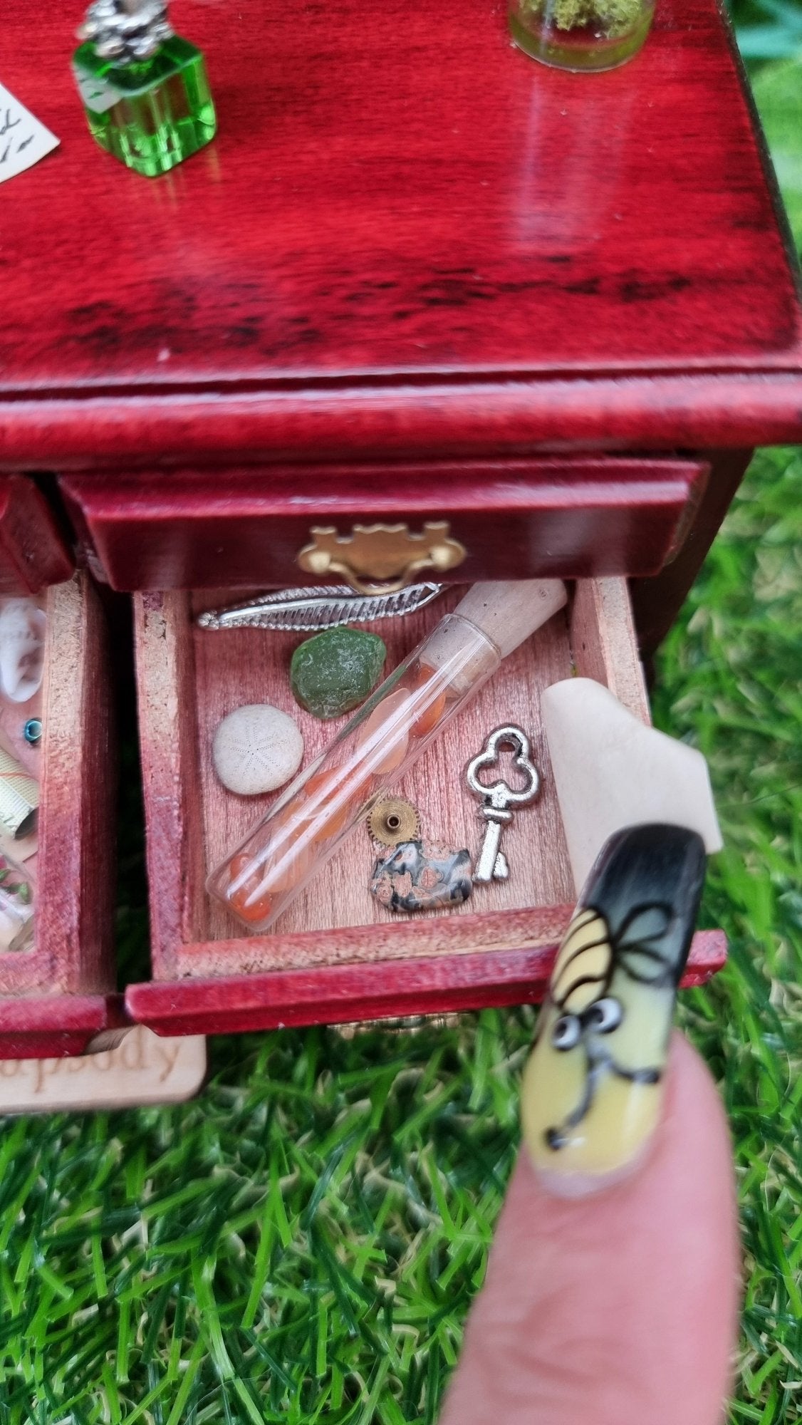 Miniature 1:12 scale dollhouse sideboard showing an open drawer containing bottle, map, parchment, trinkets and curios. The countertop has an open book and parchments. A finger holds back a parchment to reveal a hidden key