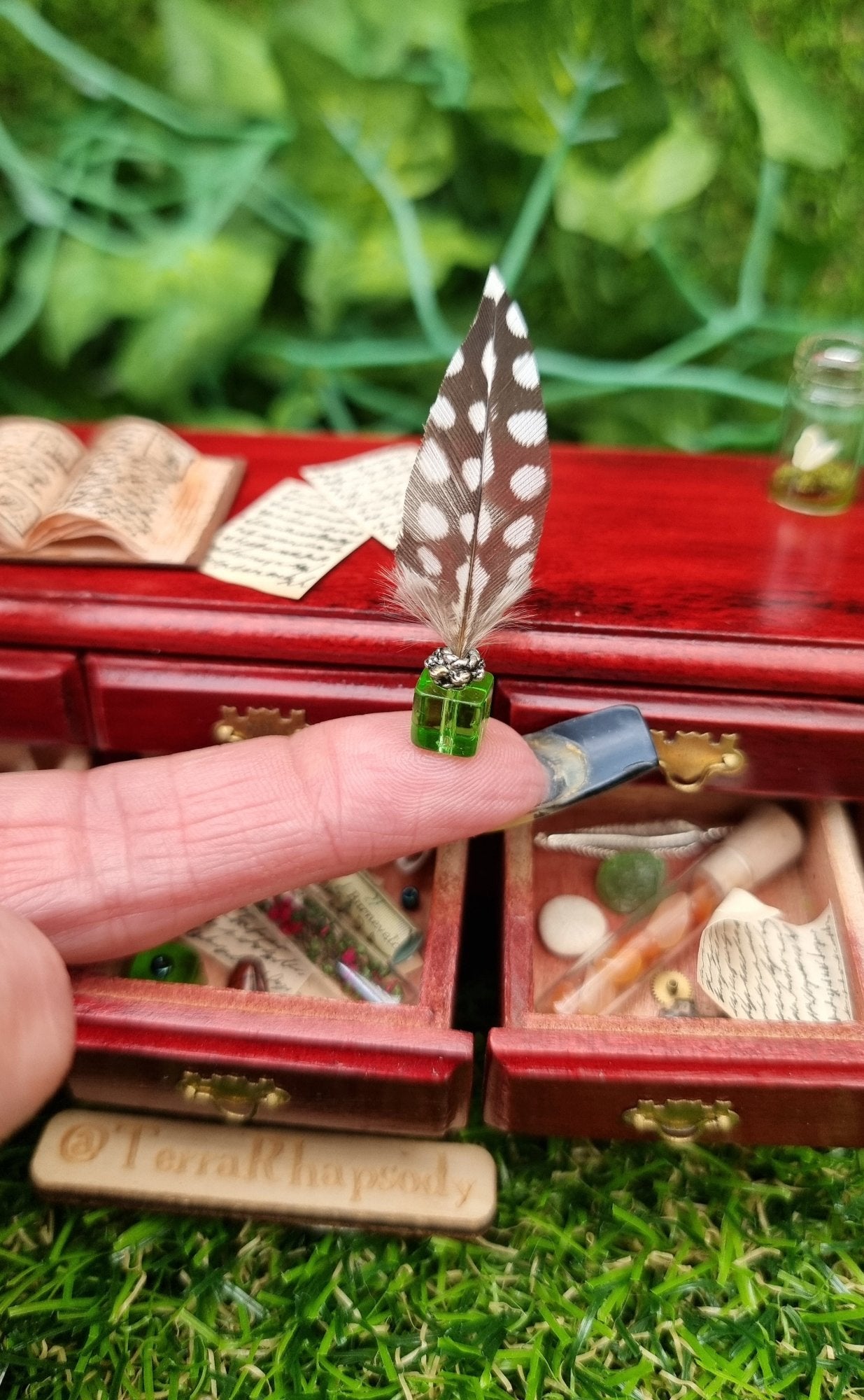 Miniature 1:12 scale dollhouse inkwell and quill sit on a finger in front of a 1:12 sideboard with 3 open drawers containing bottles, parchments, maps, trinkets and curios. The countertop has an open book and parchments