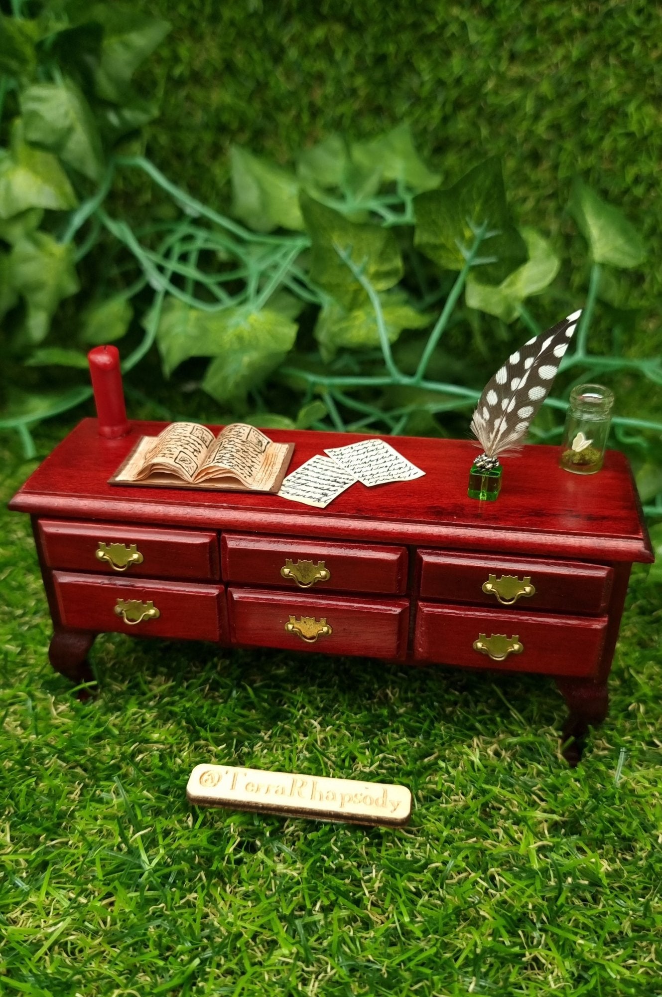 Miniature 1:12 scale dollhouse sideboard sits on a hand. There are 6 closed drawers and a countertop with a red candle, open book, butterfly bottle, quill, inkwell and parchments