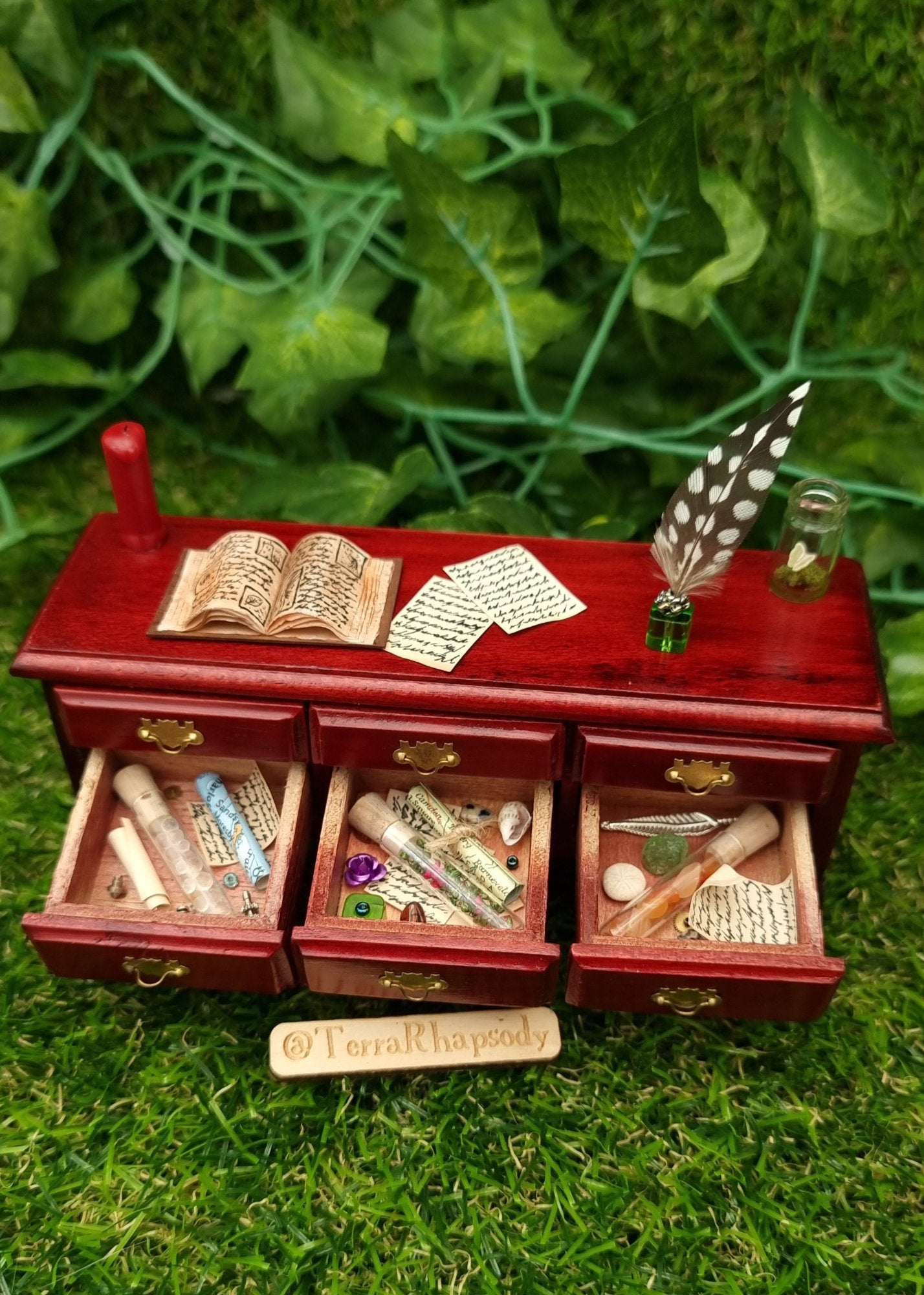 Miniature 1:12 scale dollhouse side board seen from above with 3 open drawers containing bottles, parchments, maps, trinkets and curios. The countertop has a red candle, open book, butterfly bottle, quill, inkwell and parchments