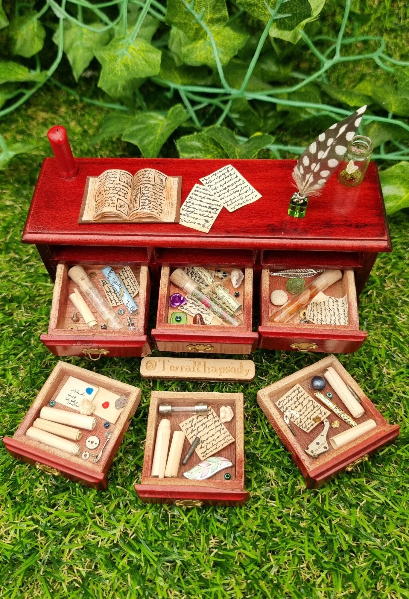 Miniature 1:12 scale dollhouse side board seen from above with 3 open drawers containing bottles parchments and curios. 3 drawers are laid out in front containing parchments, letters and trinkets. The countertop has a red candle, open book, butterfly bottle, quill, inkwell and parchments