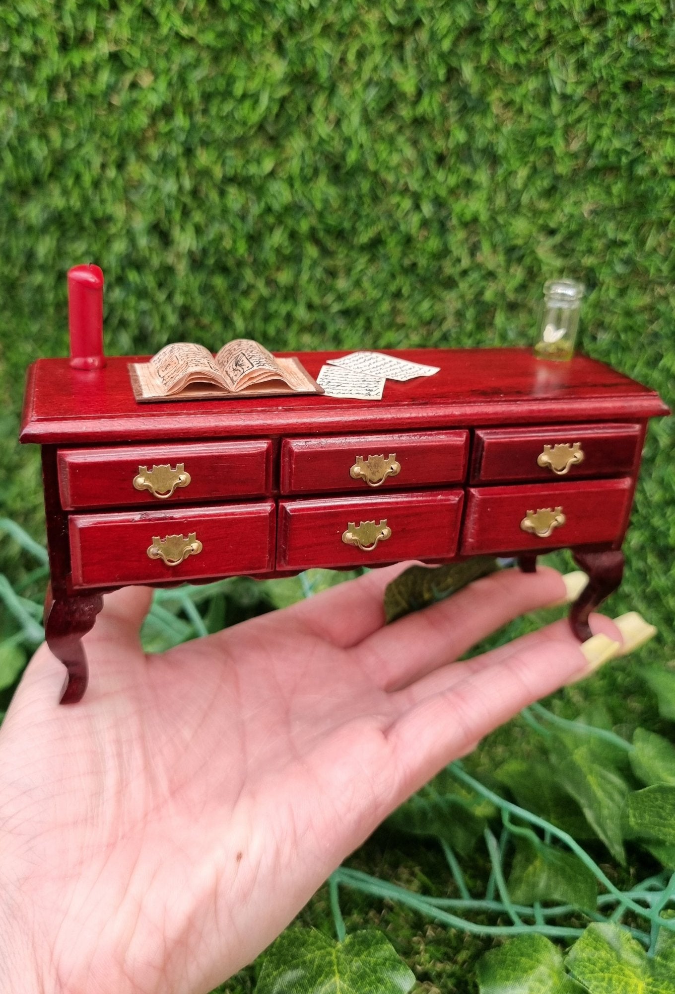 Miniature 1:12 scale dollhouse sideboard sits on a hand. There are 6 closed drawers and a countertop with a red candle, open book, butterfly bottle and parchments