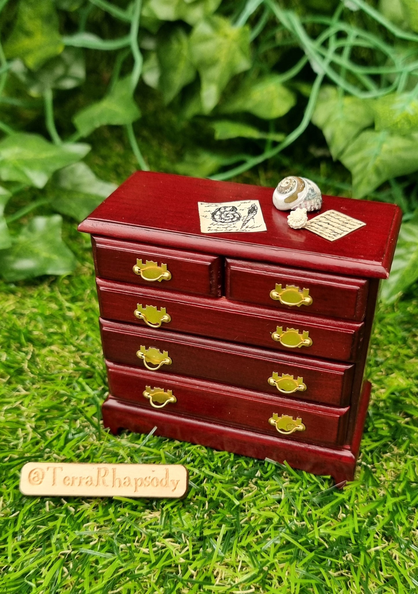 Miniature 1:12 scale chest of drawers sits on a green background. The chest is topped with shells and parchments. @terrarhapsody is written on a piece of wood beside it