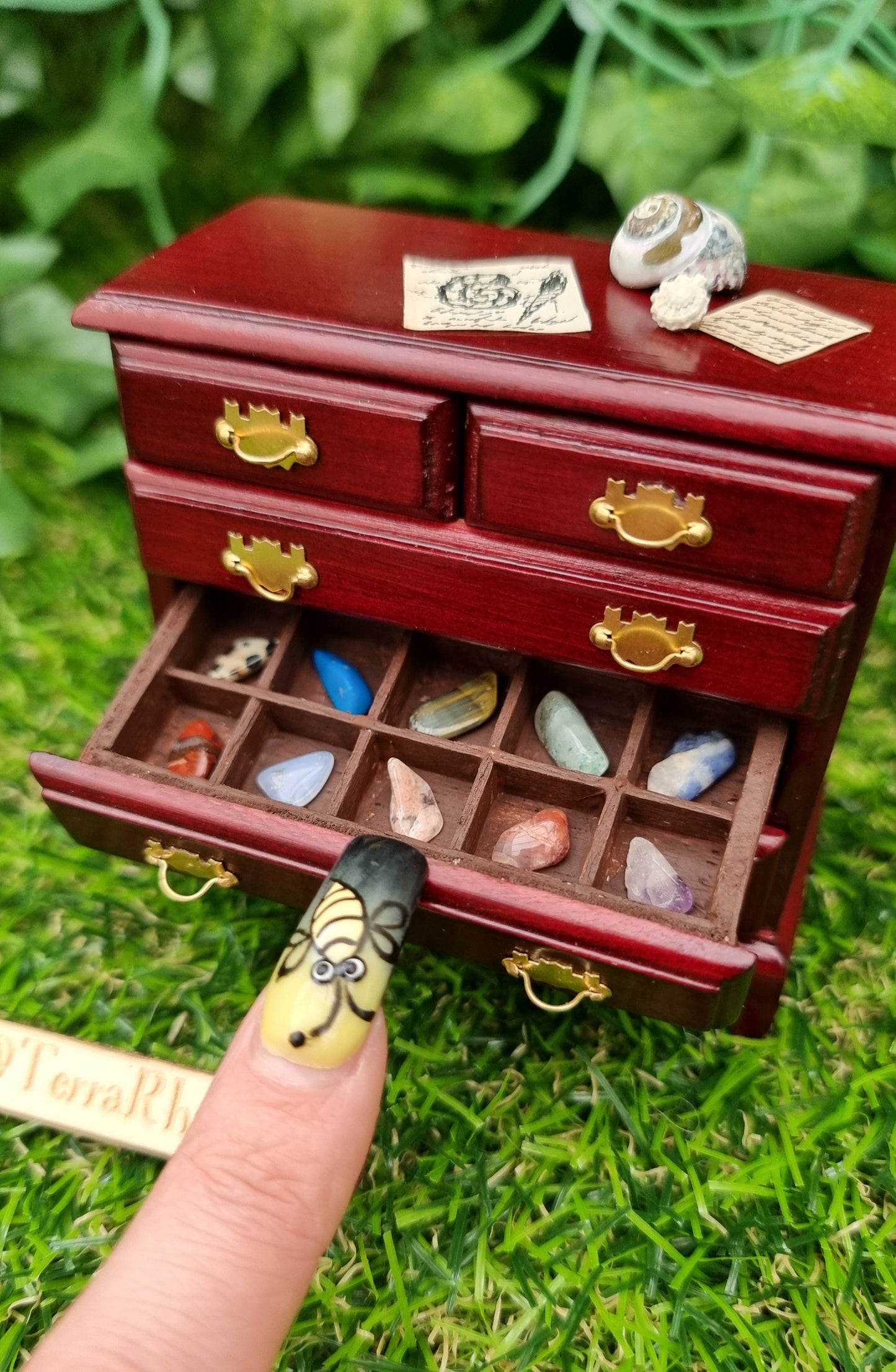 Miniature 1:12 scale chest of drawers sits on a green background. The chest is topped with shells and parchments. A finger points to one open drawer, displaying rock and mineral specimens in divided sections. @terrarhapsody is written on wood beside it