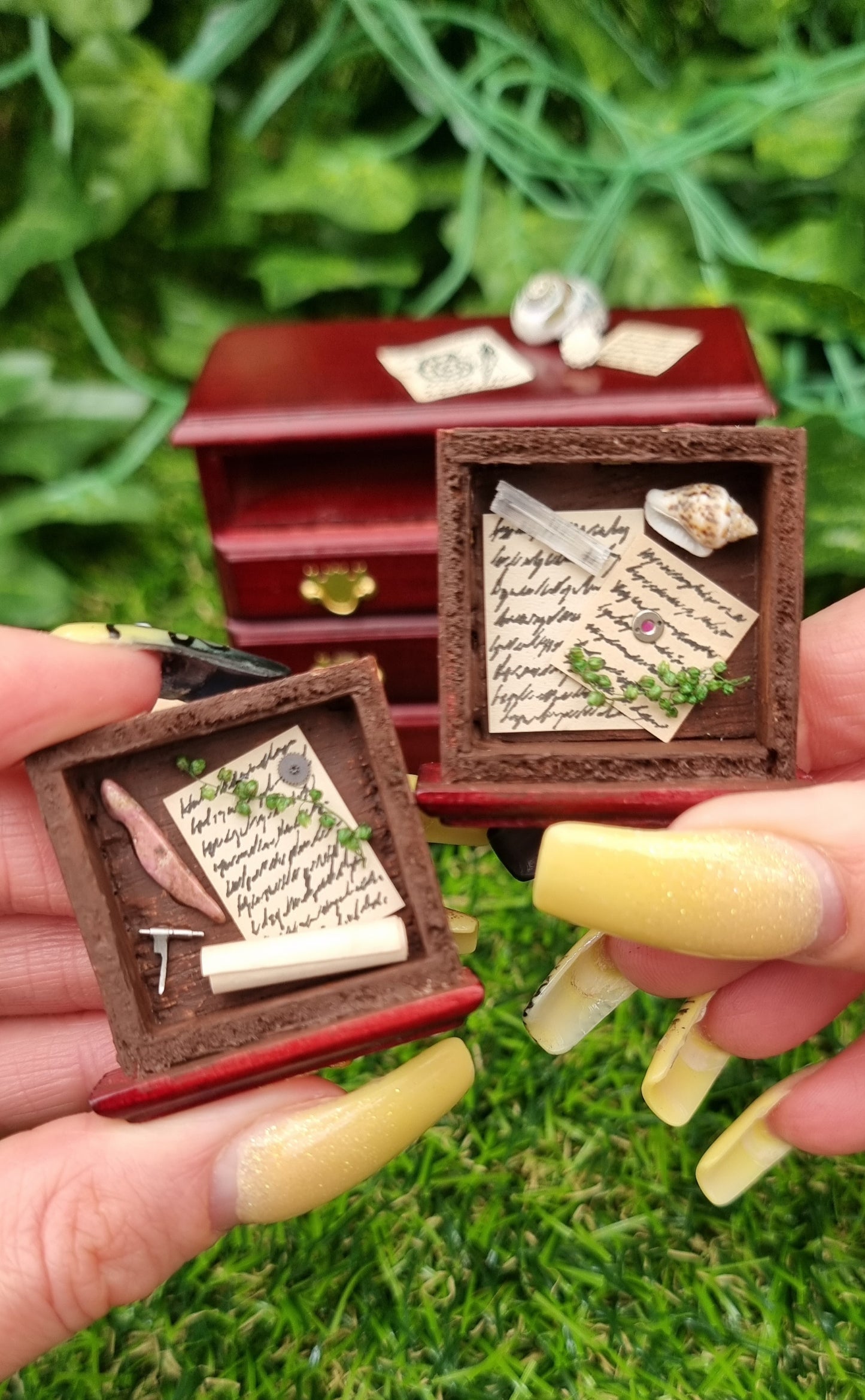 Miniature 1:12 scale chest of drawers sits on a green background. The chest is topped with shells and parchments. Fingers hold 2 drawers in front, containing parchments and curios.