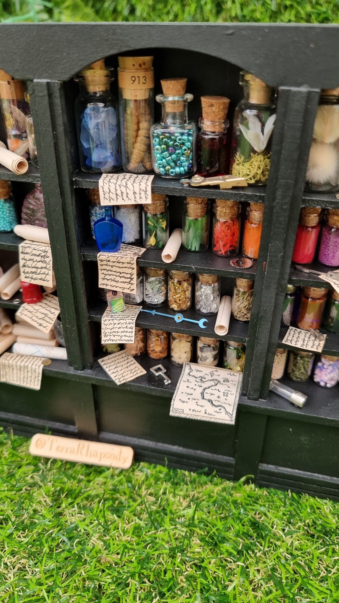 Detail of a 1:12 scale black shelving unit. A third of the unit is shown, with 4 shelves full of glass bottles containing various different potion ingredients. Written parchments sit on some of the shelves beneath various vessels and curios. There is a map and key on the bottom shelf. @TerraRhapsody is written on a piece of wood in front of it