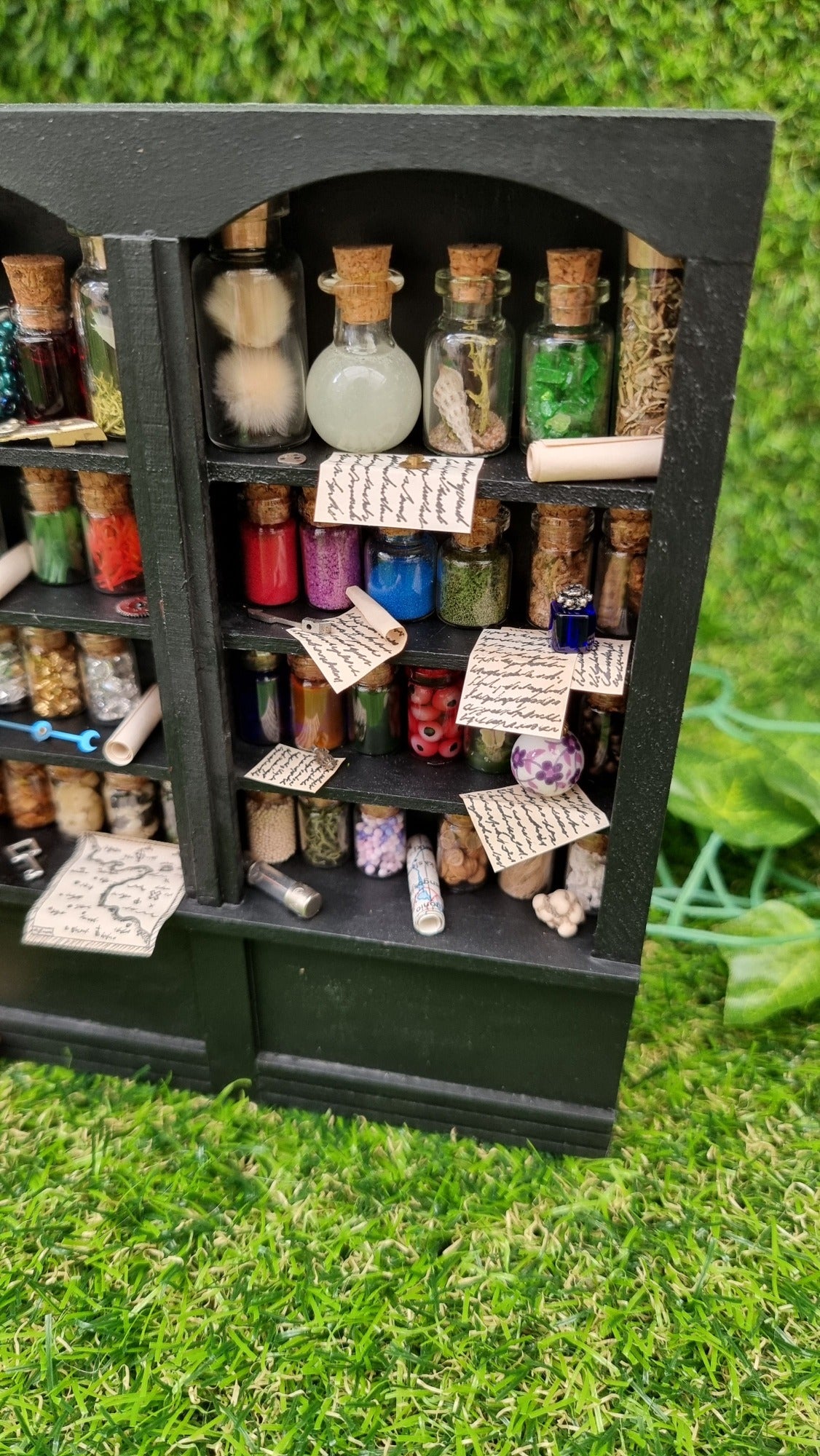Detail of a 1:12 scale black shelving unit. A third of the unit is shown, with 4 shelves full of glass bottles containing various different potion ingredients. Written parchments sit on some of the shelves beneath various vessels and curios