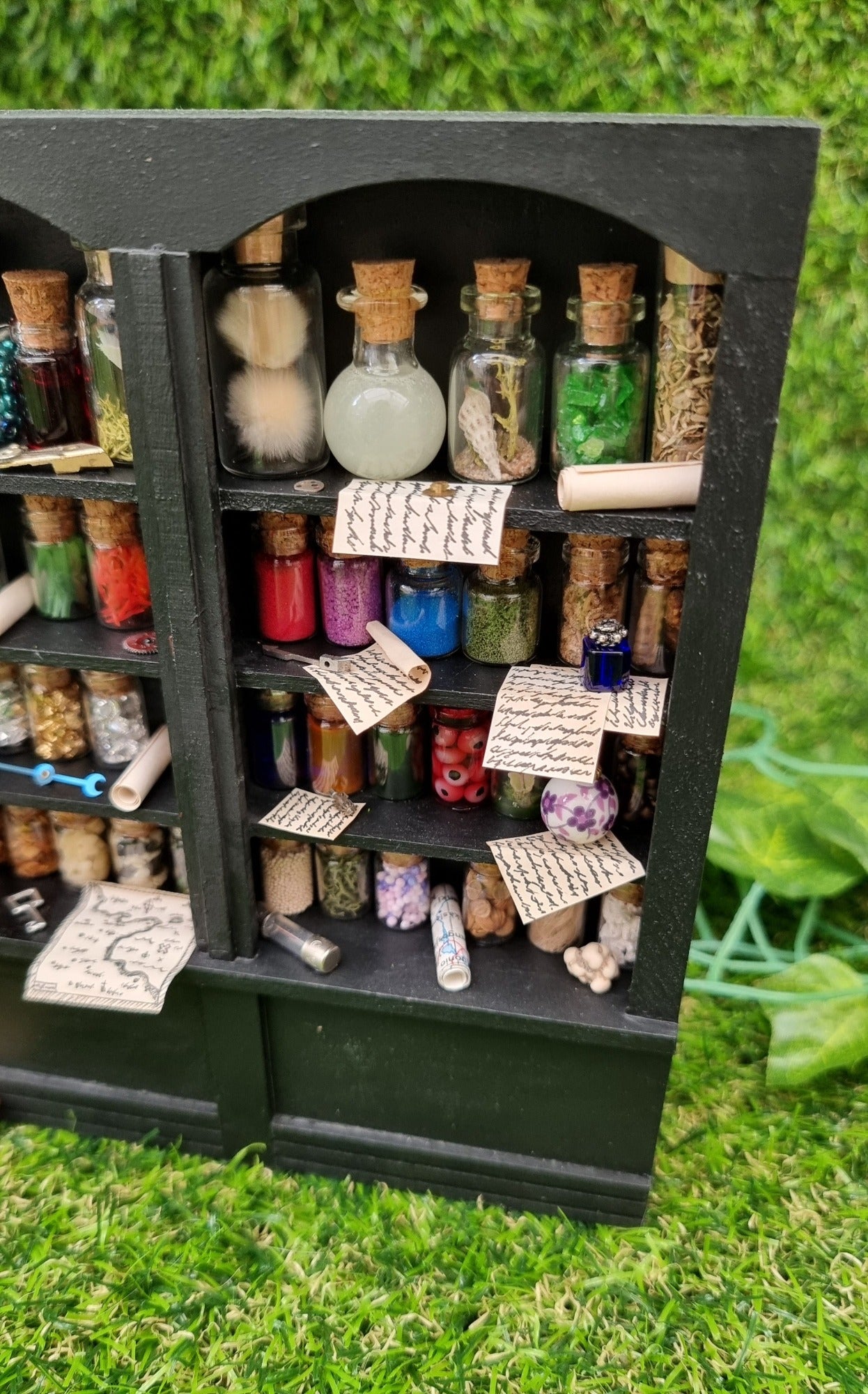 Detail of a 1:12 scale black shelving unit. A third of the unit is shown, with 4 shelves full of glass bottles containing various different potion ingredients. Written parchments sit on some of the shelves beneath various vessels and curios
