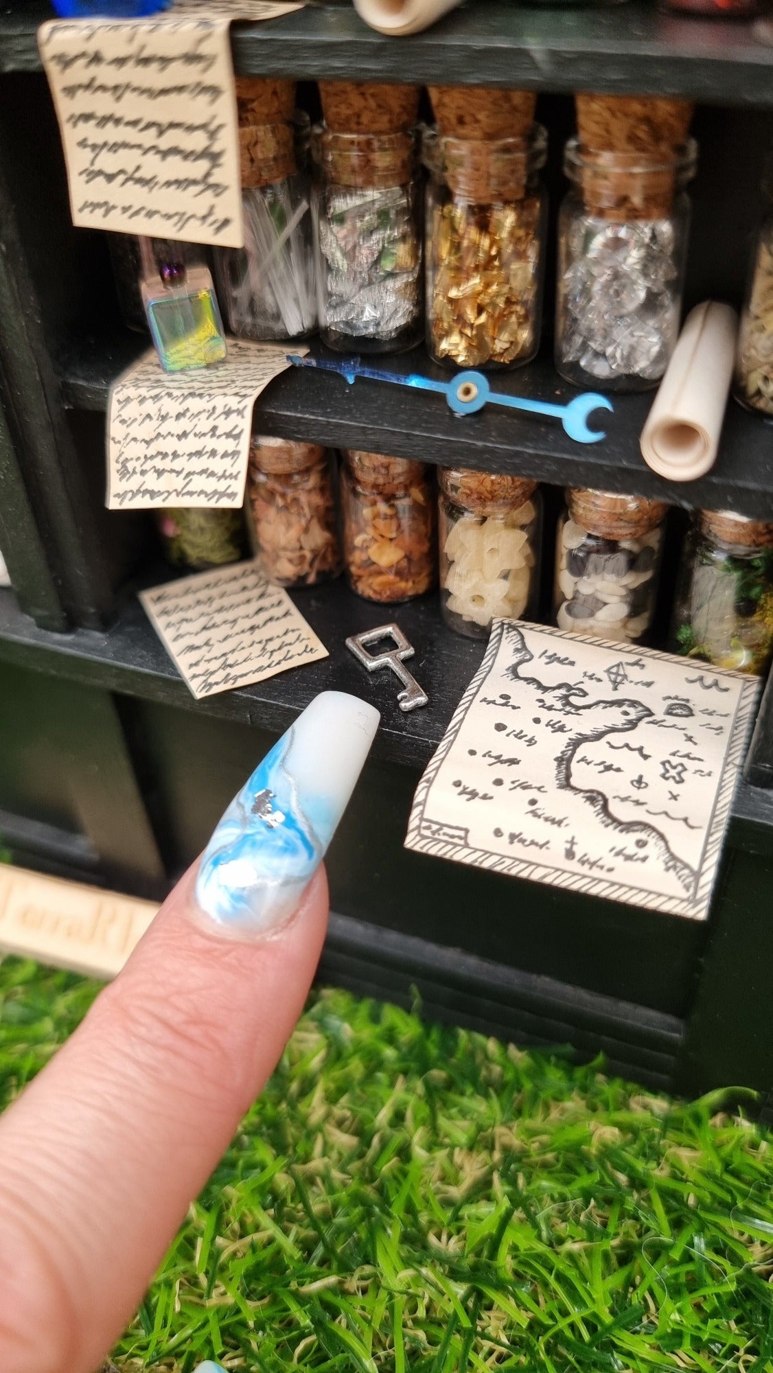 Detail of a 1:12 scale black shelving unit. Two shelves are shown, full of glass bottles containing various different potion ingredients. Written parchments sit on some of the shelves beneath various vessels and curios. A finger points at a key on the bottom shelf besides a map