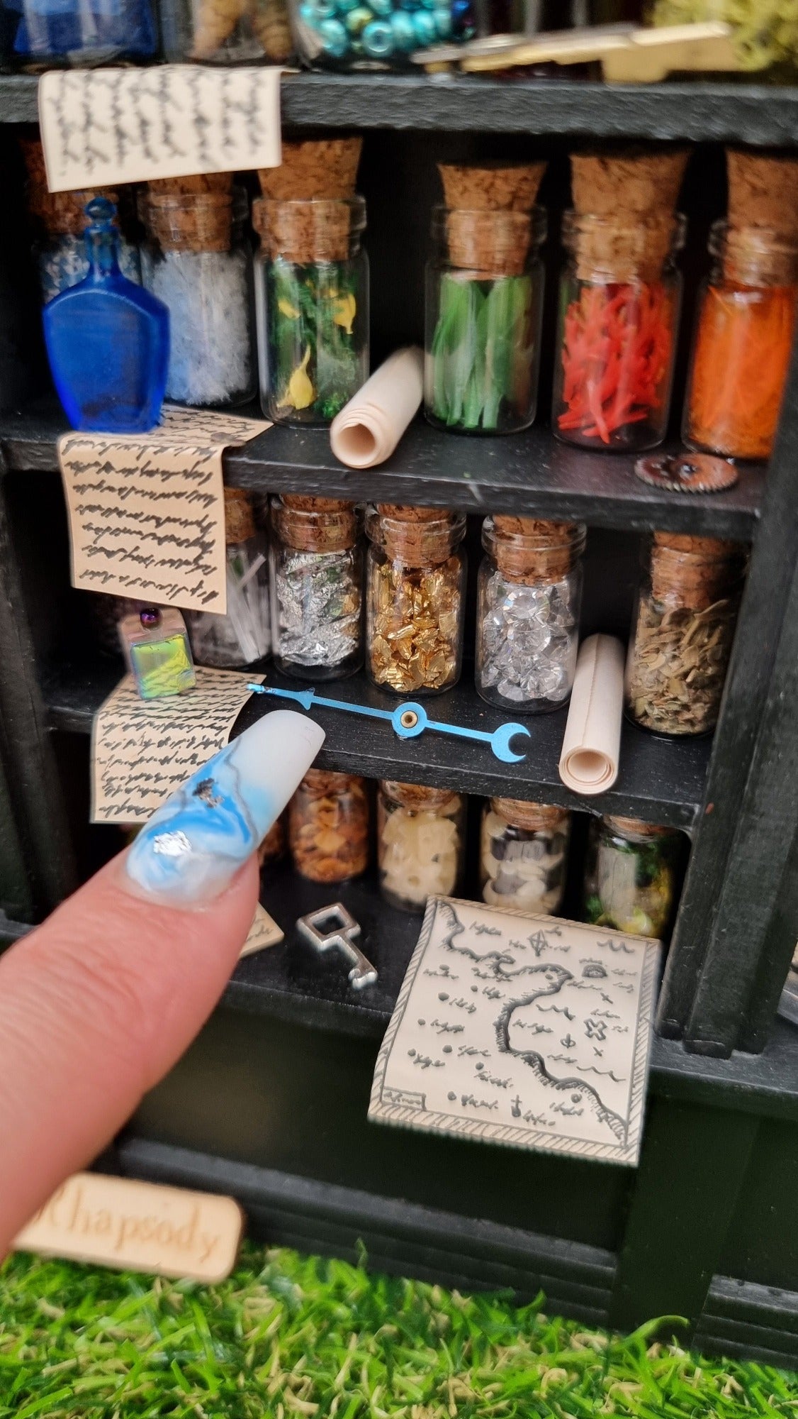 Detail of a 1:12 scale black shelving unit. A third of the unit is shown, with 3 shelves full of glass bottles containing various different potion ingredients and written parchments. There are various vessels, curios, map and key on the bottom shelf. A finger points at a blue metal dial