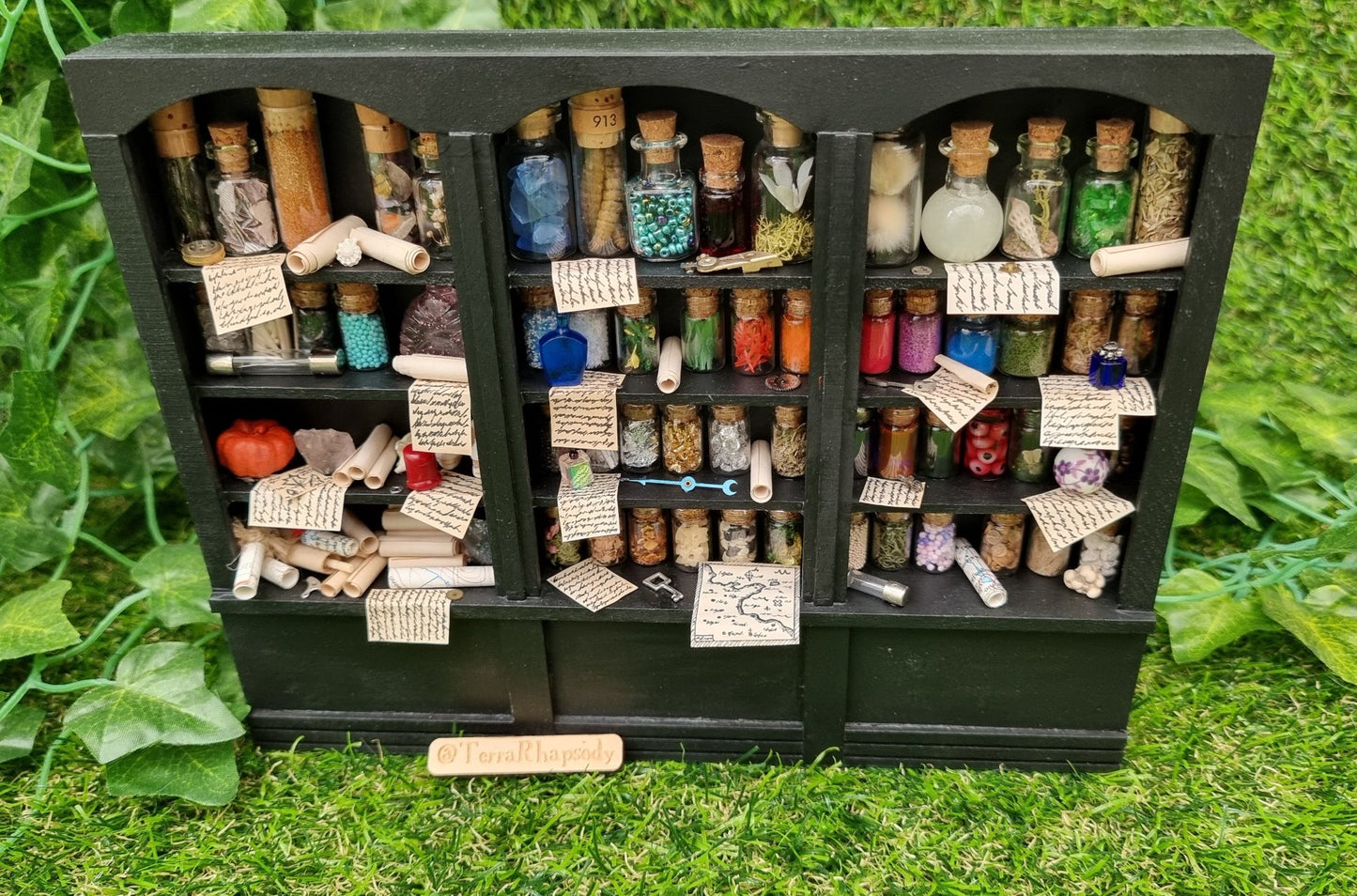 Miniature 1:12 scale black shelving unit sits against a green background. Each shelf is filled with glass vials full of potion ingredients, with other small vessels and parchments. 2 shelves have curios and trinkets, including skulls and parchment rolls