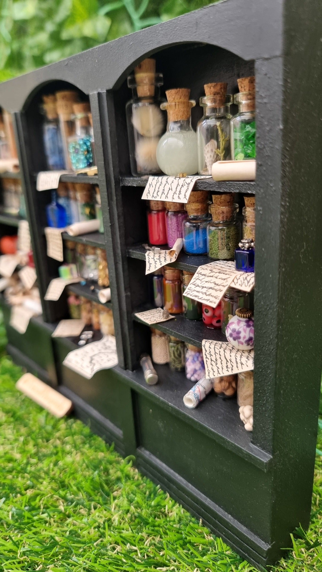 Detail of a 1:12 scale black shelving unit viewed from the side. A third of the unit is shown, with 4 shelves full of glass bottles containing various different potion ingredients. Written parchments sit on some of the shelves beneath various vessels and curios