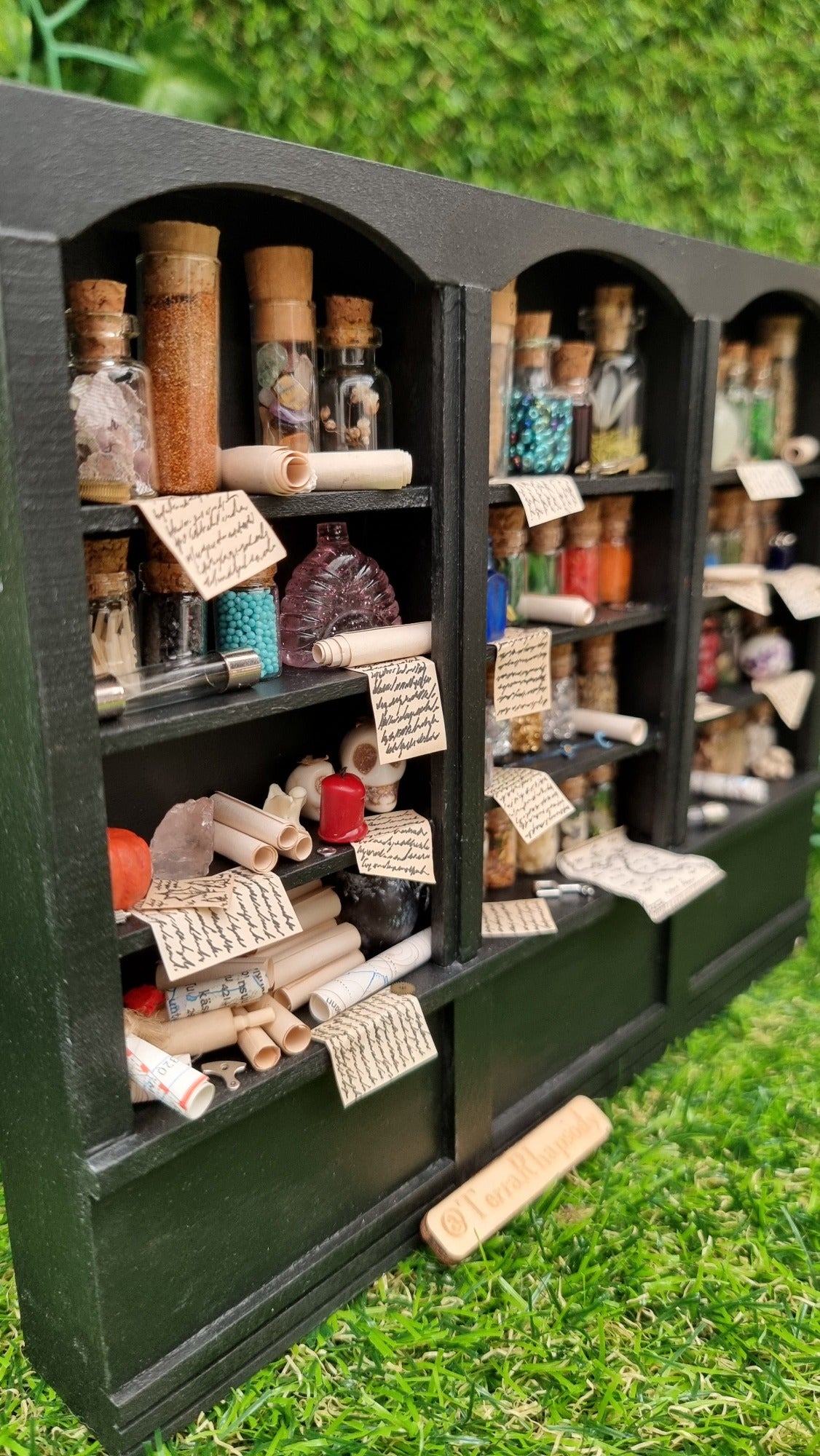 Miniature 1:12 scale black shelving unit shown from one end sits against a green background. Each shelf is filled with glass vials full of potion ingredients, with other small vessels and parchments. 2 shelves have curios and trinkets, including skulls and parchment rolls