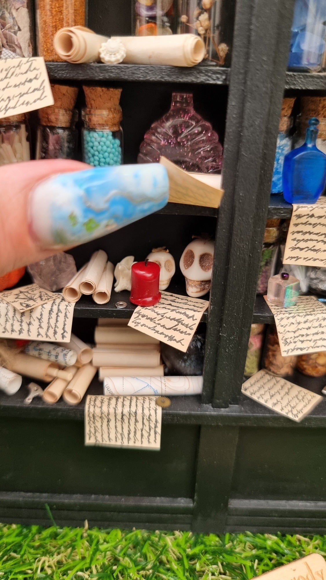 Detail of a 1:12 scale black shelving unit. A thumb lifts a parchment revealing two skulls and a red candle. Filled glass bottles can be seen on the shelves above, parchment and map rolls on the shelf below