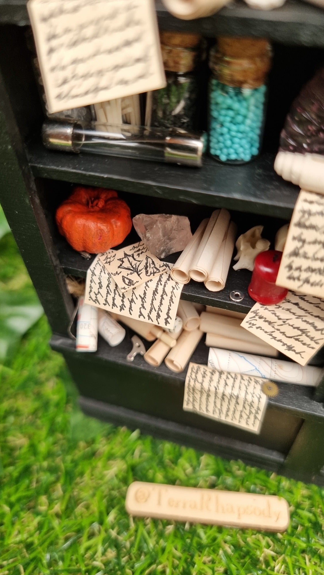 Looking down from the top shelf of a 1:12 scale black shelving unit showing details of parchment rolls, pumpkin, candle and written parchmet notes with diagrams. @TerraRhapsody is written on a piece of wood in front of it