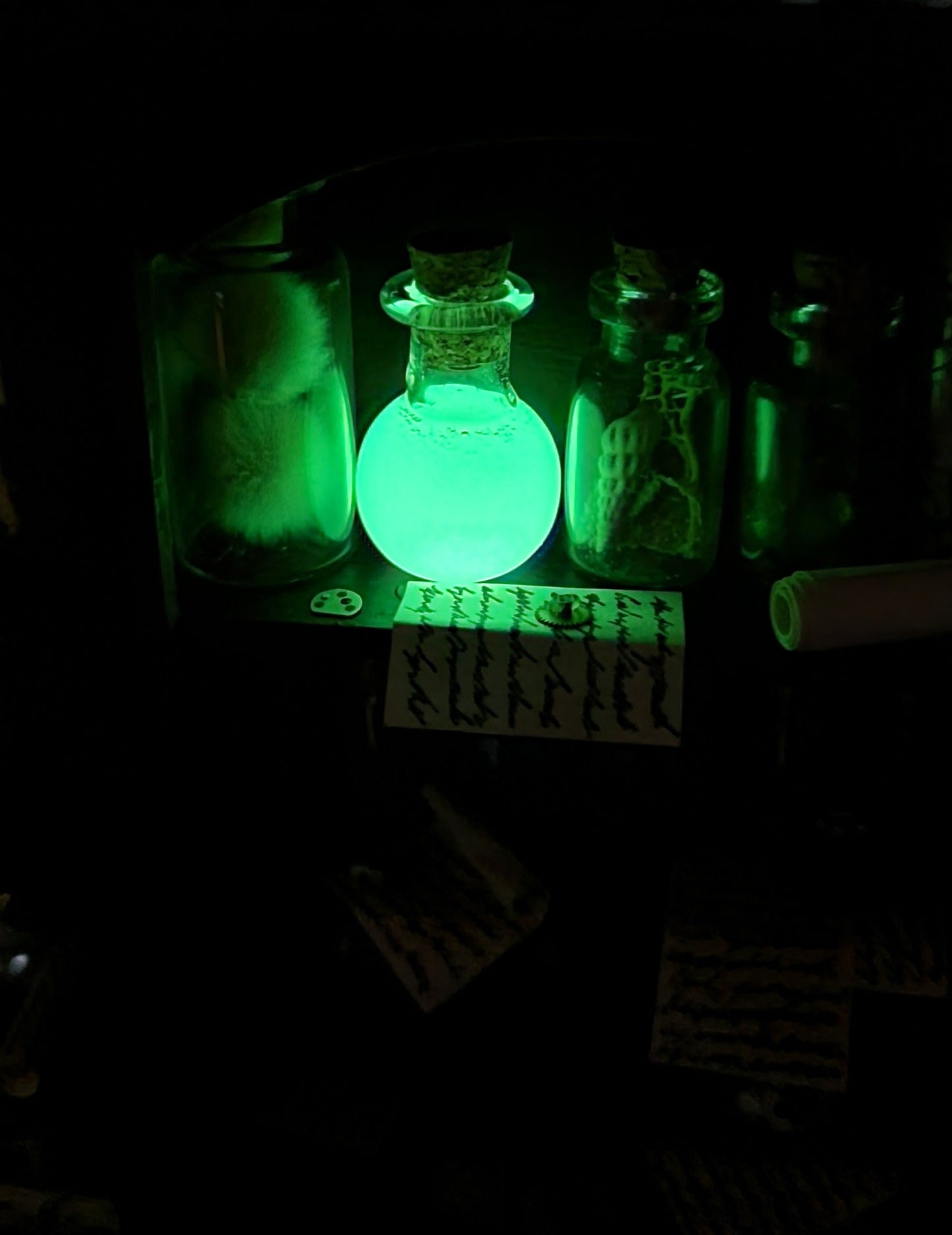 A 1:12 scale dollhouse shelf is shown in darkness as a boiling flask shaped glass vial glows green in the dark, illuminating surrounding glass bottles and a parchment note.