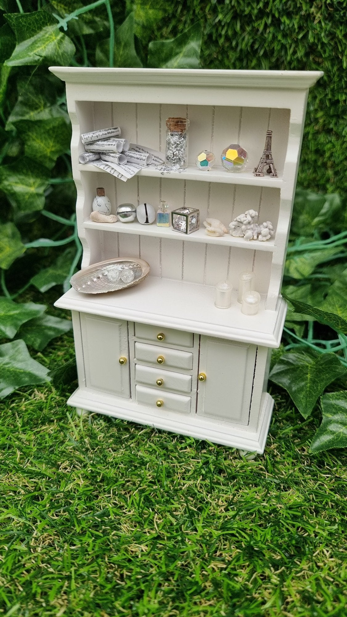 Miniature 1:12 scale white dresser sits against a green background. The dresser has closed cupboard doors and 4 closed drawers. The counter top and 2 shelves above contain candles, crystals, glass bottle, shells, coral and sheet music