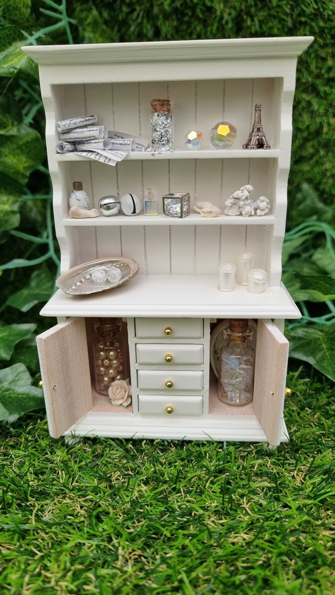 Miniature 1:12 scale white dresser sits against a green background. The dresser has open cupboard doors revealing a white rose and bottles of gemstones. There are 4 closed drawers. The counter top and 2 shelves above contain more candles, crystals, glass bottle, shells, coral and sheet music