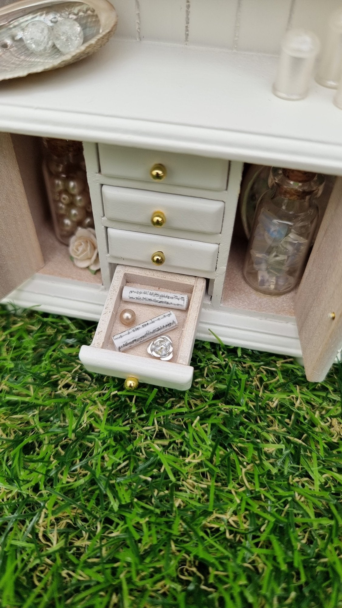 Miniature 1:12 scale white dresser sits against a green background. The dresser has open cupboard doors revealing a white rose and bottles of gemstones. 1 out of 4 drawers is open, revealing sheet music, silver metal rose and cream pearl. The counter top above has candles and a large shell with crackle glass spheres